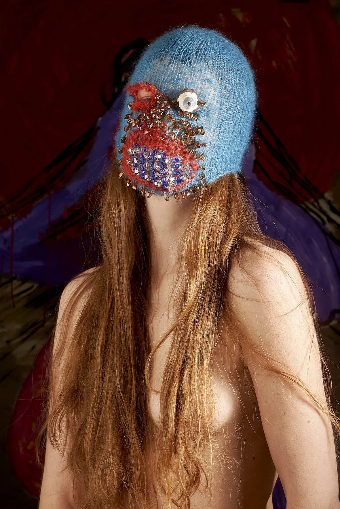 Person with long hair wearing a blue, beaded knit mask with red and metallic accents, against a colorful abstract background.
