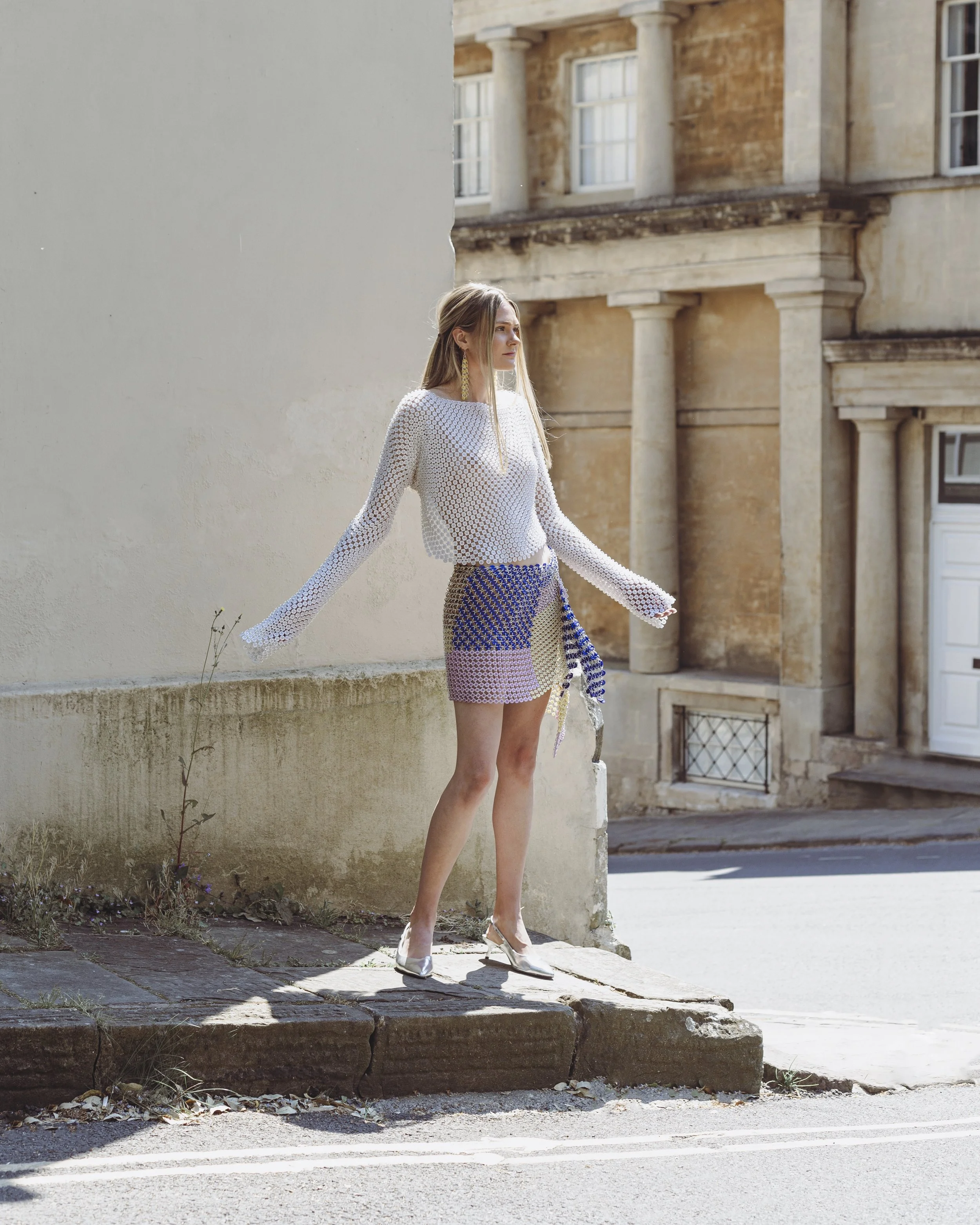 A person standing on a sidewalk near a building, wearing a white knit sweater and a colorful skirt, with hands outstretched. The background features an old stone structure.