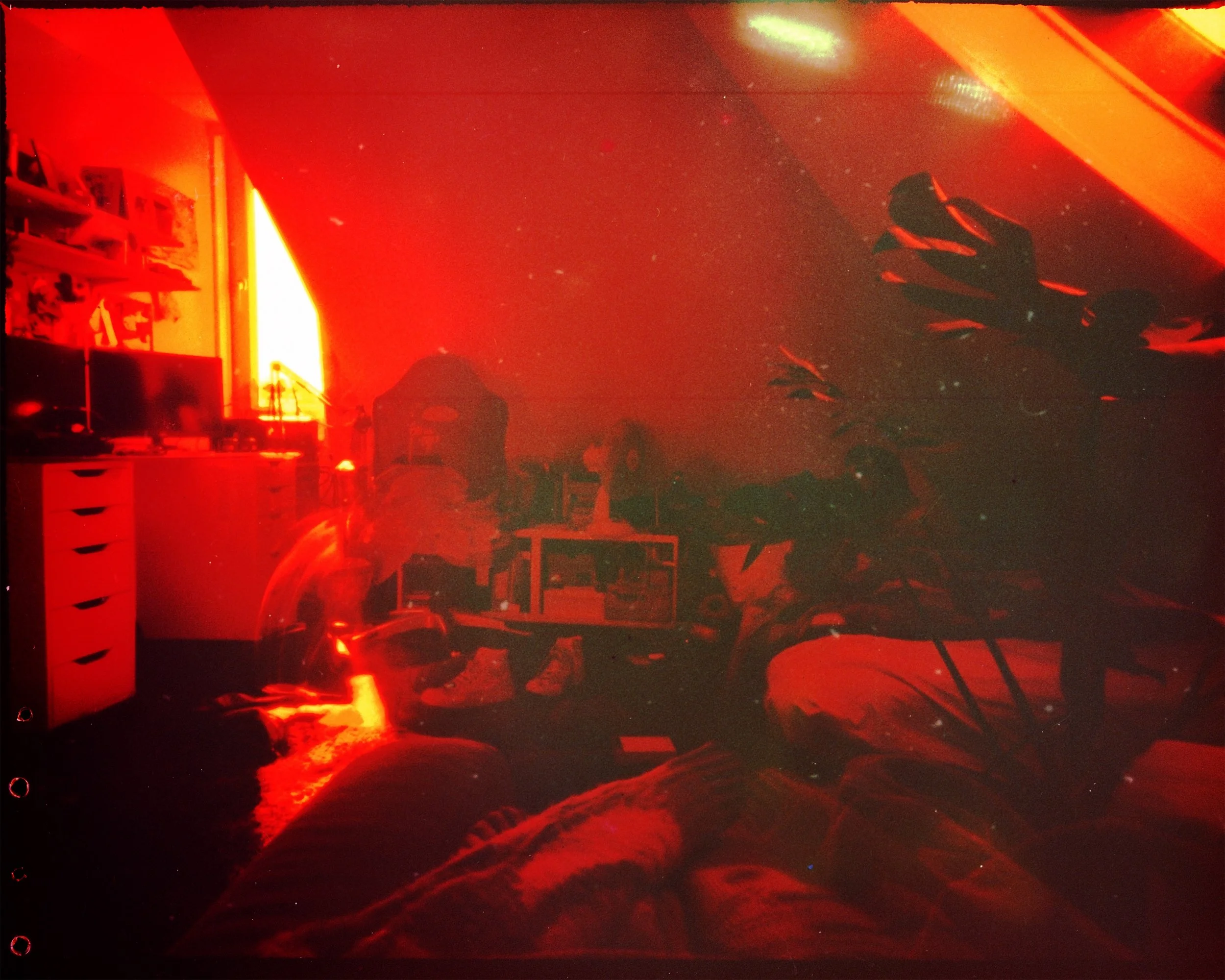 A dimly lit room with red lighting, featuring a desk with a computer monitor and drawers, a chair, and shelves. A large plant is visible, and personal items are scattered, including shoes and decorations. The atmosphere is cozy with a vintage or moody aesthetic.