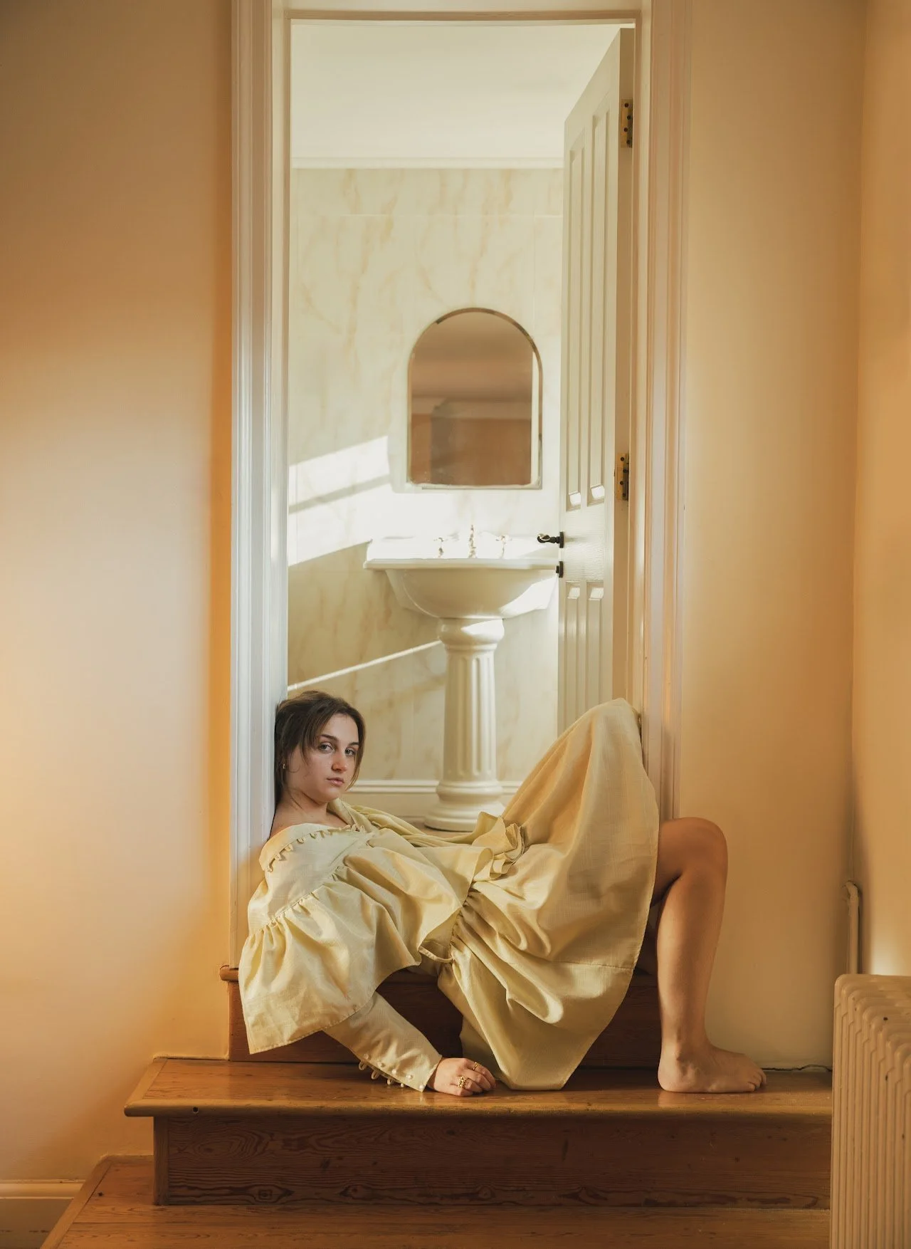 Woman in beige dress sitting on a staircase near an open door with a bathroom visible, featuring a sink and mirror.