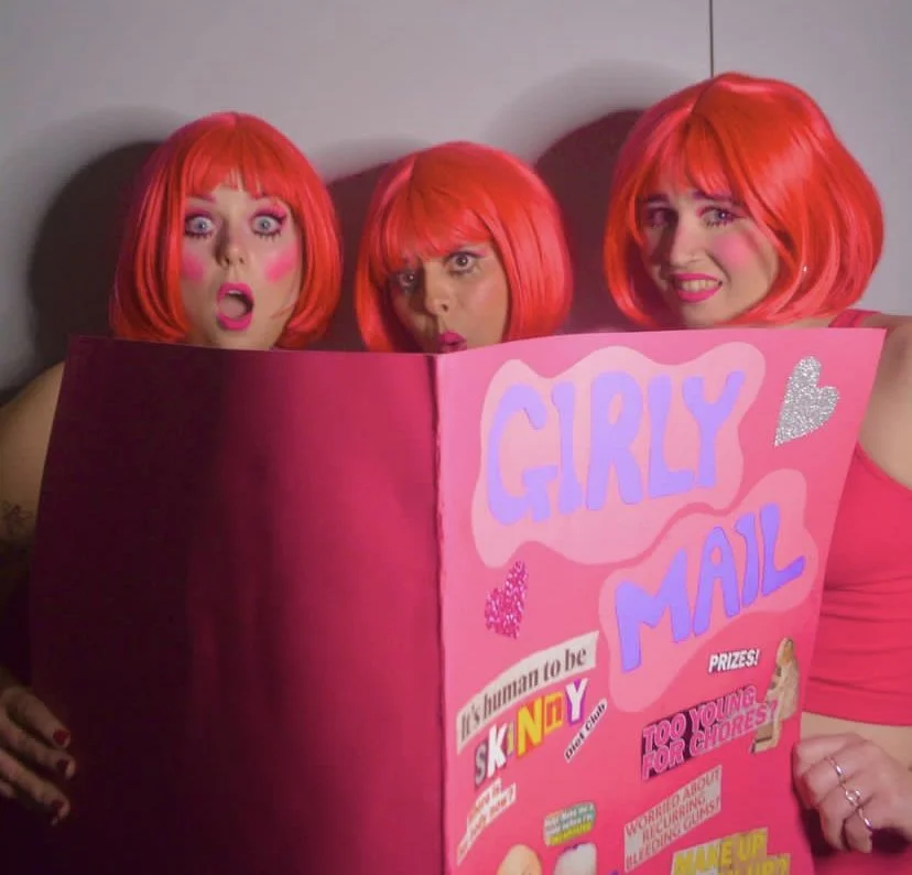 Three people wearing bright pink wigs and makeup, holding and looking at a large pink sign titled 'Girly Mail' with various stickers and decorations.