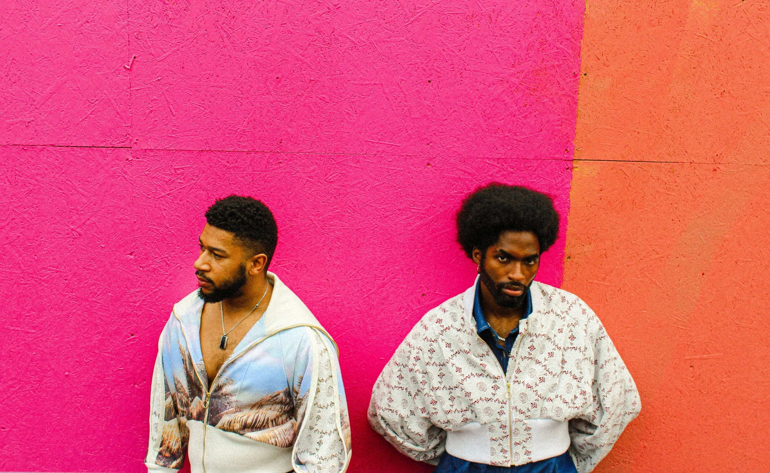 Two men standing against a vibrant pink and red wall, wearing colorful jackets and looking in different directions.