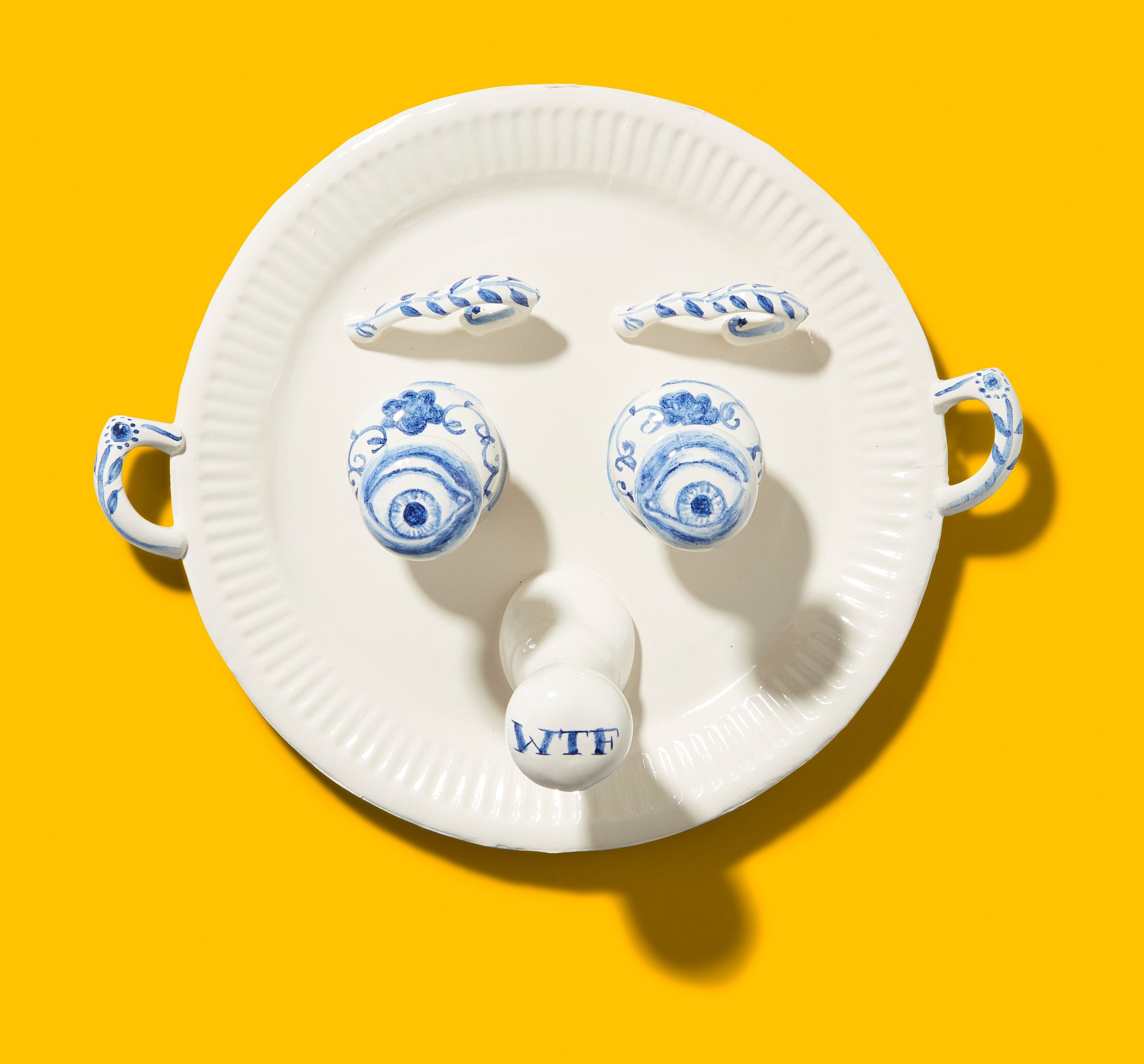 Plate with handles, arranged to resemble a face, with decorative blue designs and the letters "WTF" on a yellow background.