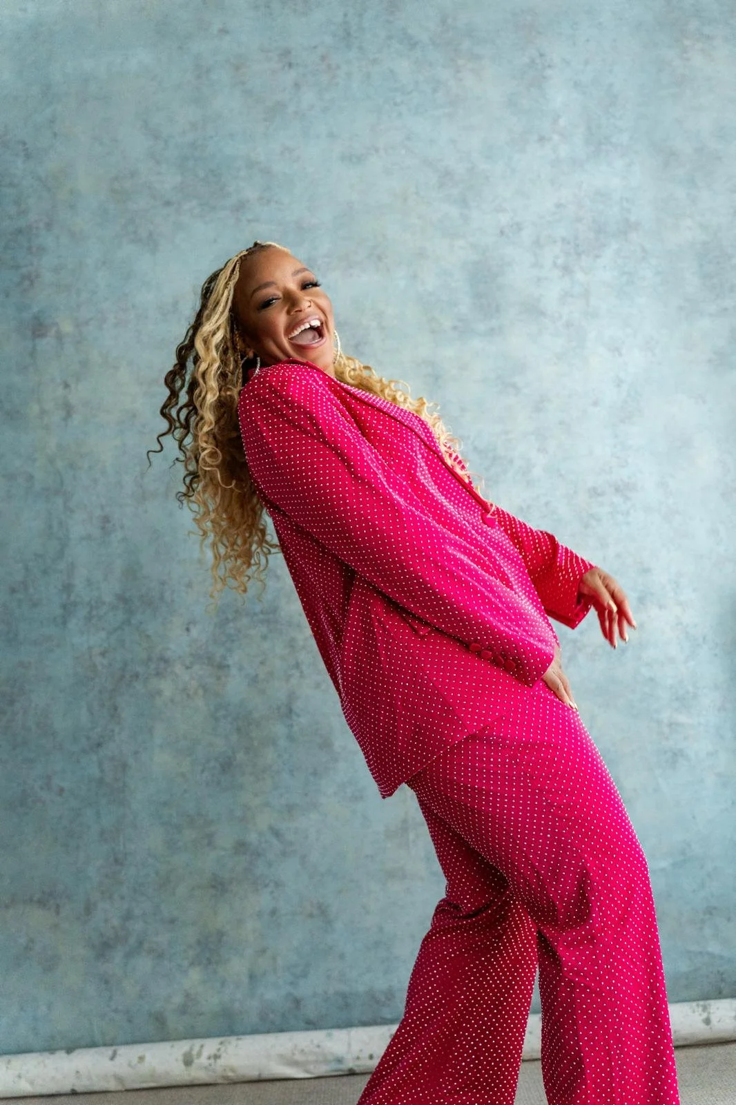 Marisha Wallace in a pink polka dot suit laughing against a blue background.