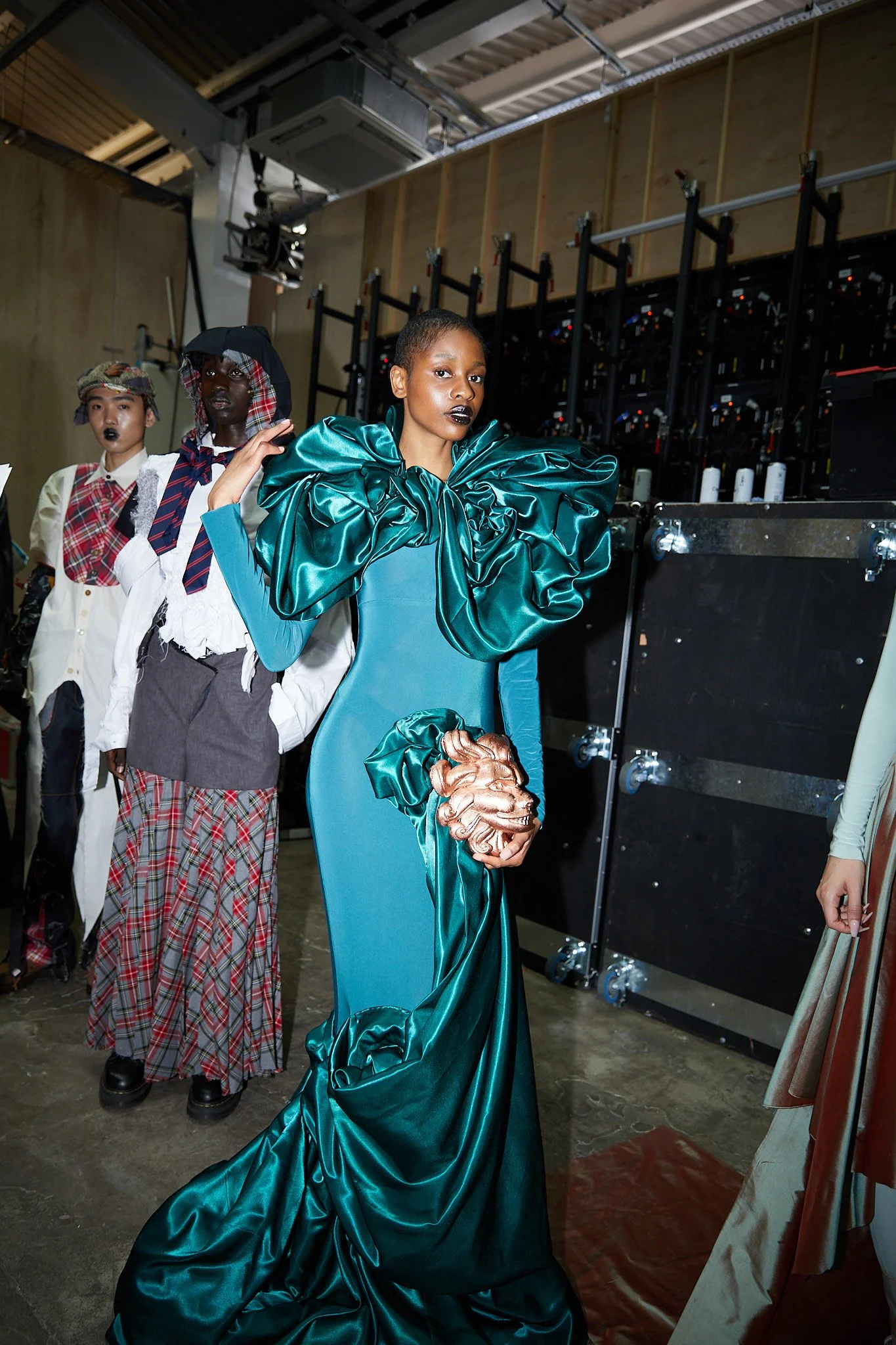 Backstage at a fashion show featuring models in avant-garde outfits. The central model wears a teal dress with bold ruffled detailing and metallic accents, surrounded by other models in unique, creative attire.