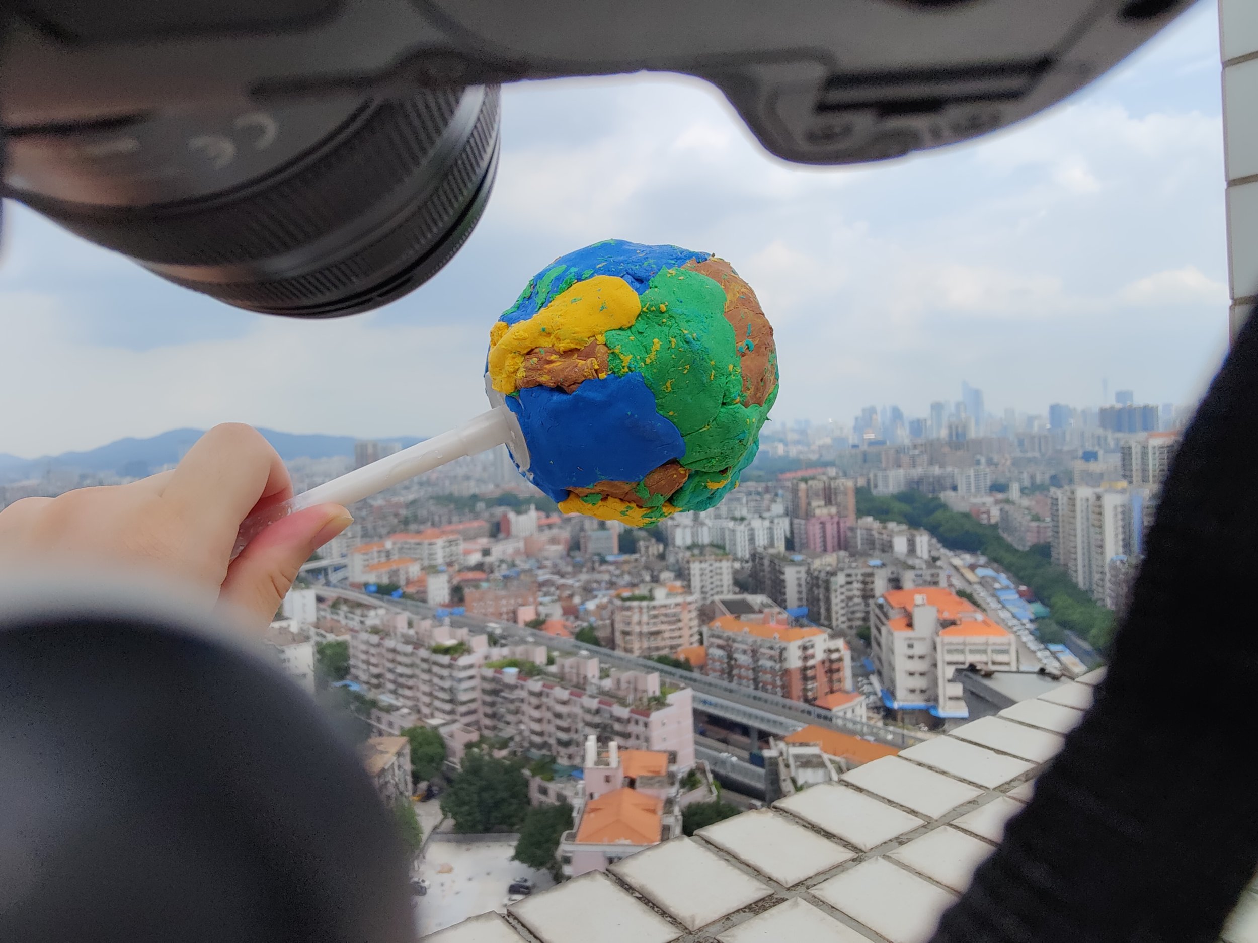 A hand holding a globe lollipop in front of a cityscape view seen through a camera lens.