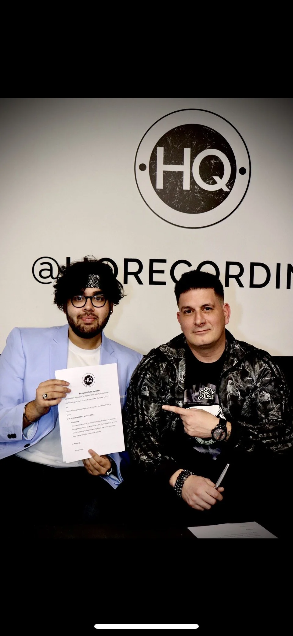 Two people holding a document, sitting in front of a wall with HQ logo. The person on the left wears glasses and a blue jacket, holding up a paper. The person on the right is pointing at the document, wearing a dark patterned jacket.