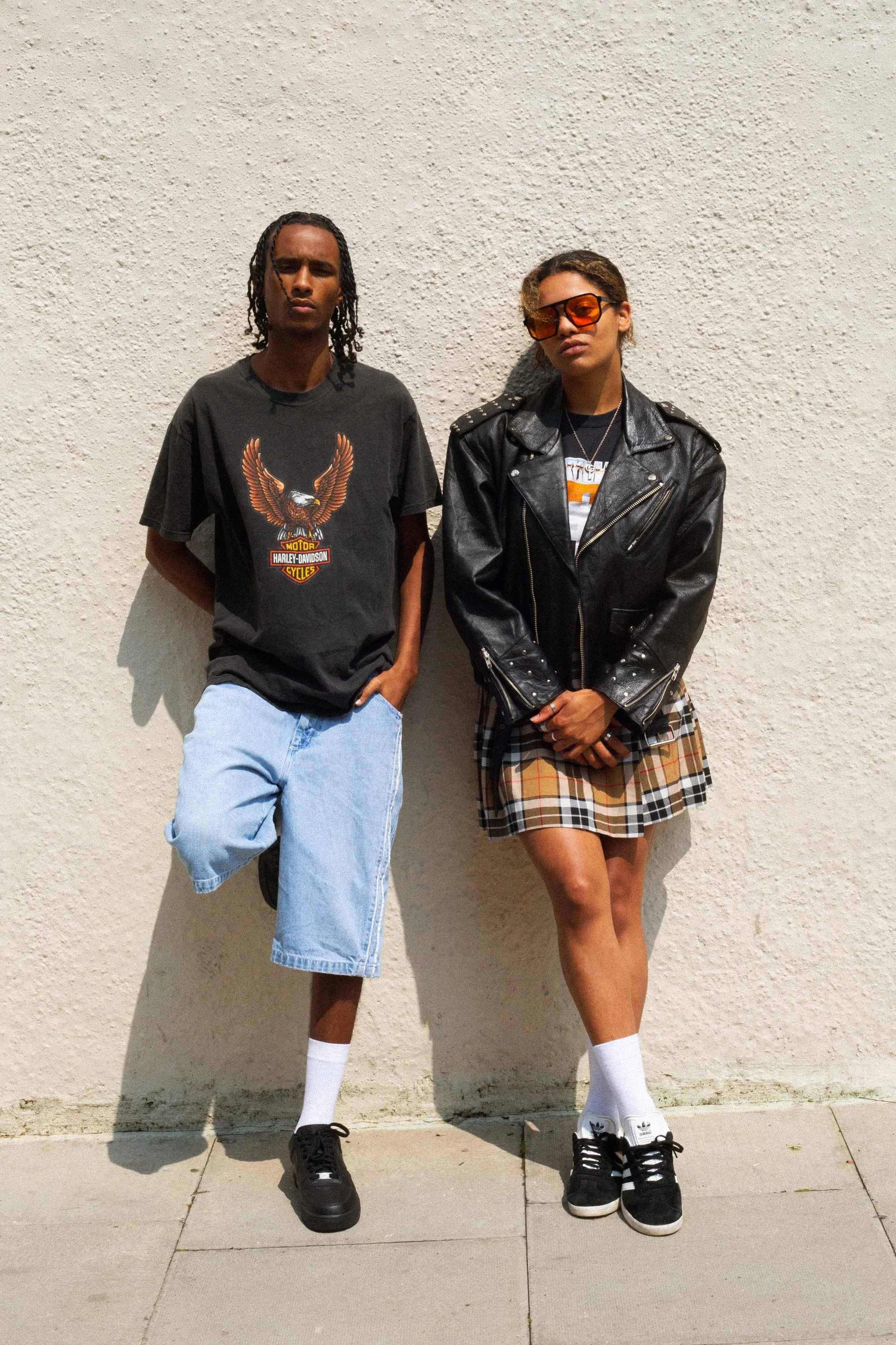 Two individuals standing against a textured wall; one wearing a black graphic T-shirt and denim shorts, the other in a black leather jacket, plaid skirt, and sunglasses.