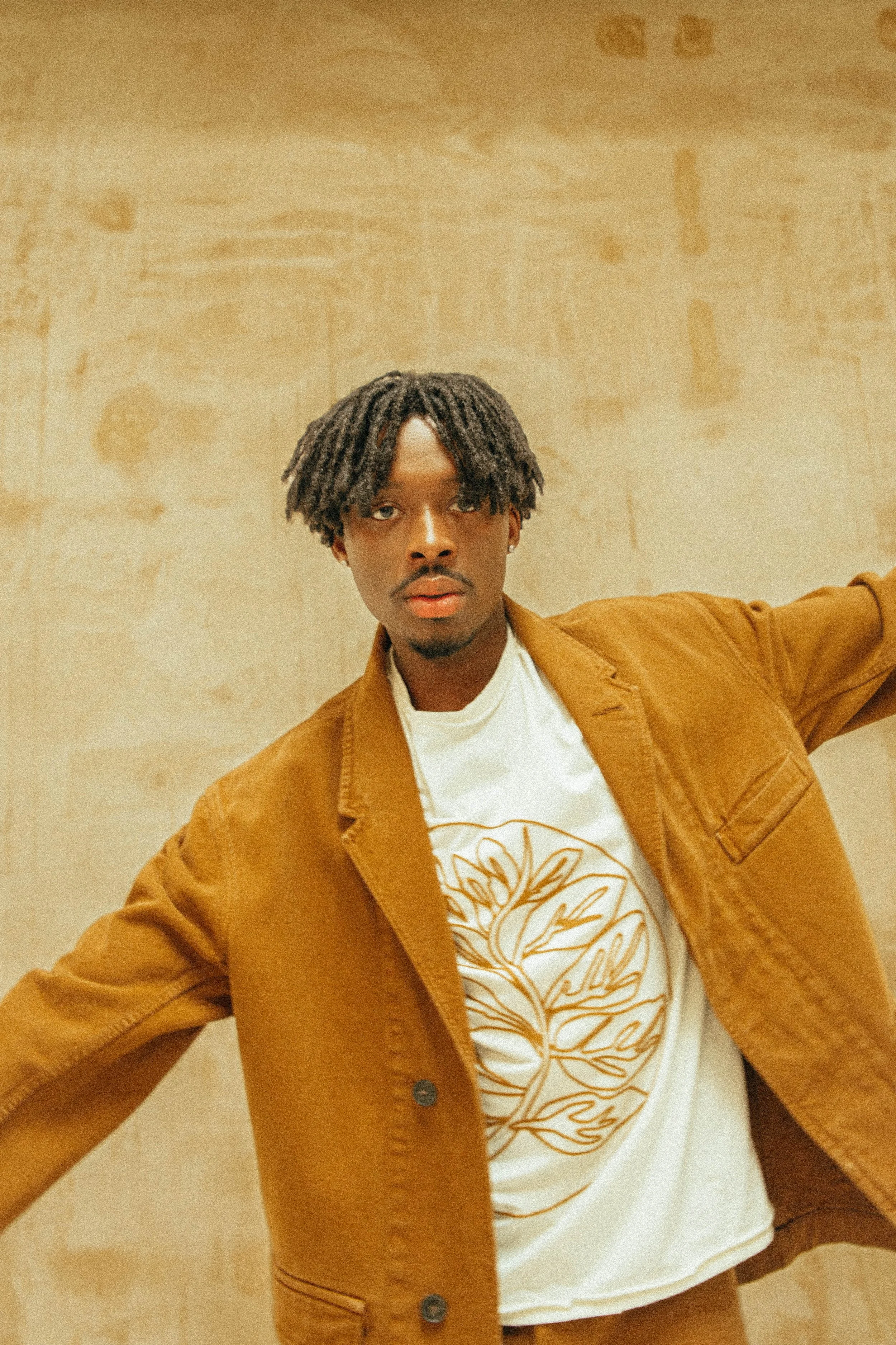 Person wearing a brown jacket and a white t-shirt with a botanical design, standing in front of a beige textured wall.