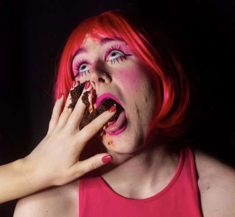 Person with pink wig and dramatic makeup being fed cake with a hand