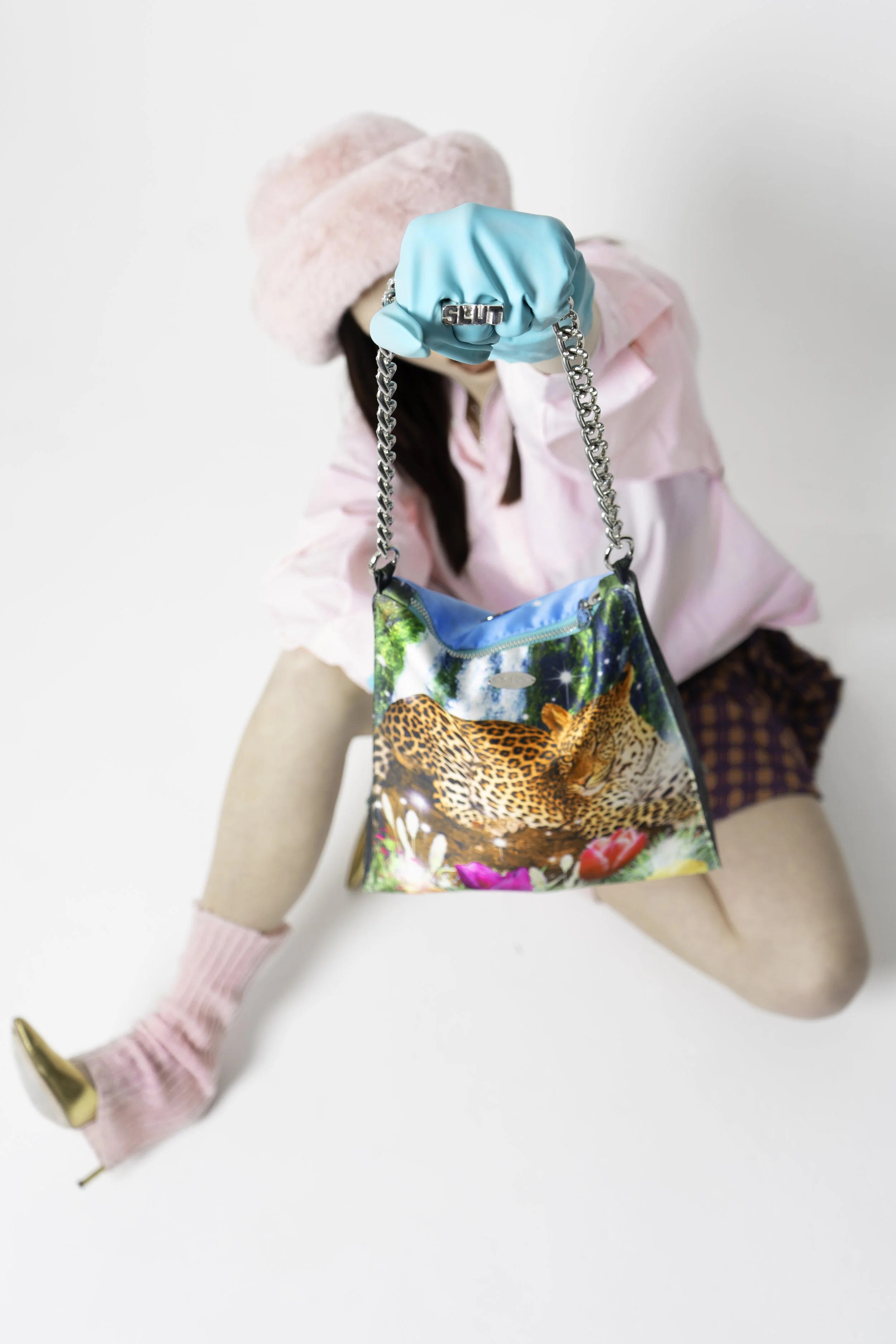 Person wearing a fluffy pink hat and pink socks with high heels, holding a colorful purse displaying a leopard image and chain strap. The person is dressed in a pink shirt and patterned shorts.