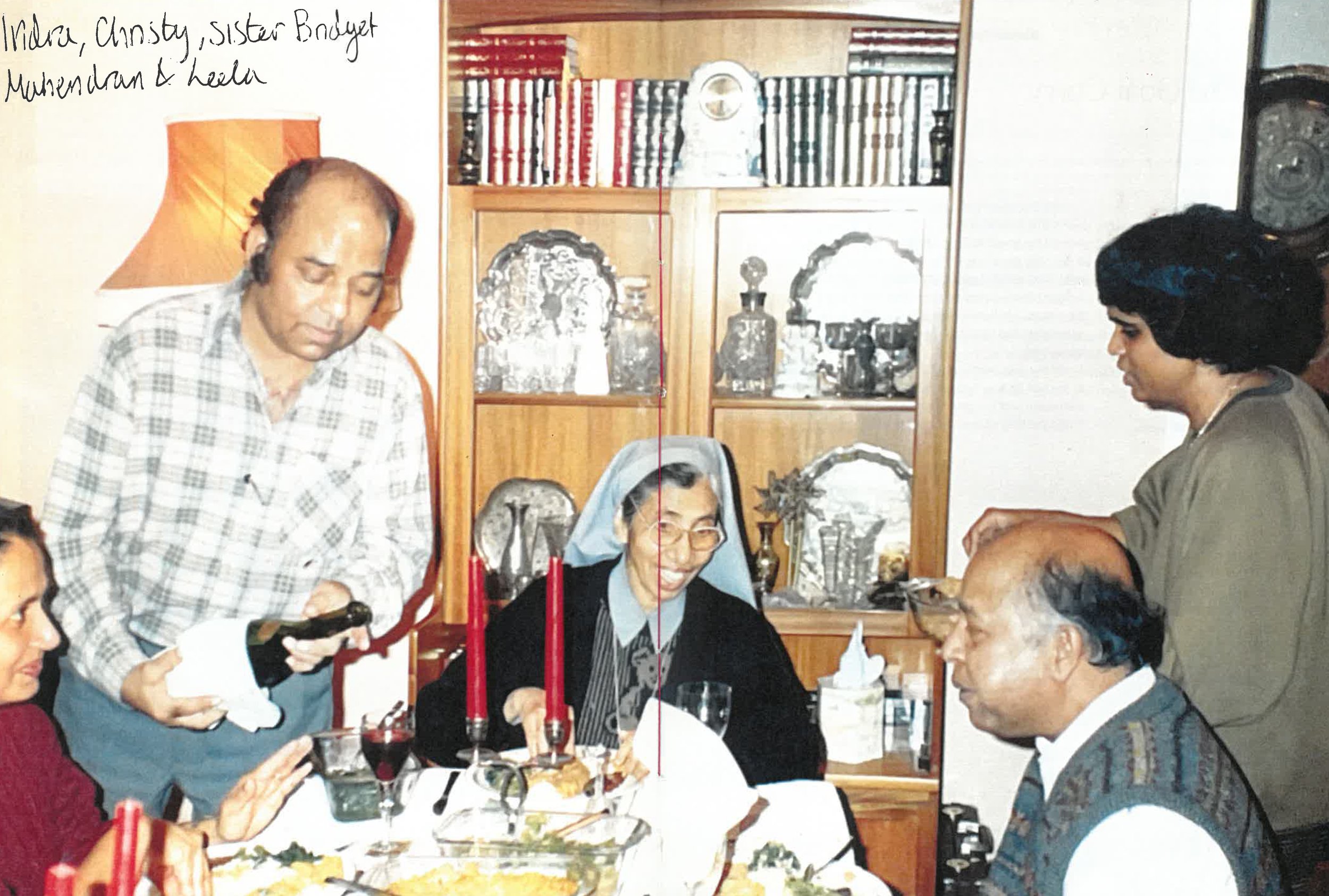 A group of people gathered around a dining table with food and candles, in front of a shelf with books and decorative items. The scene is indoors, with a lamp and handwritten names visible on one side.