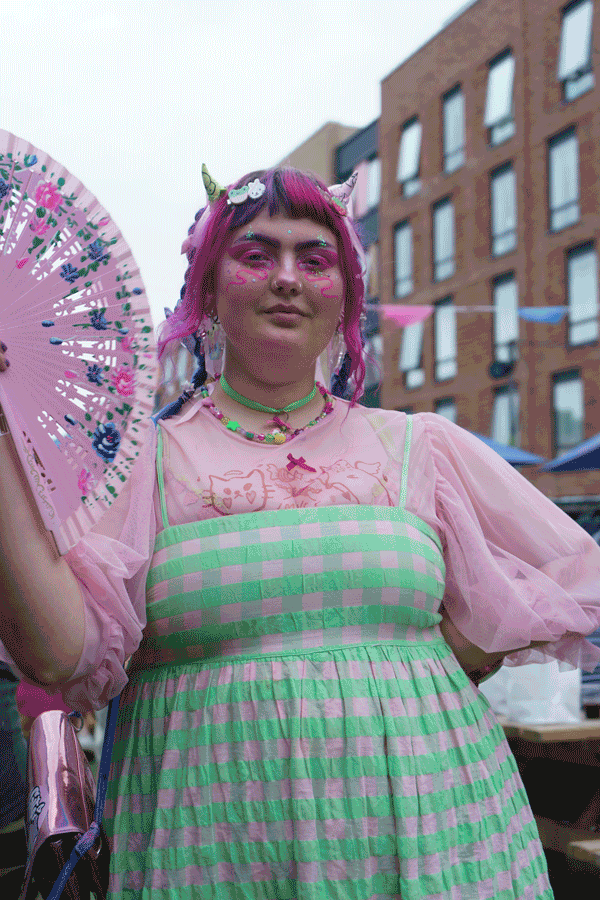 Person with pink and green clothing, vibrant hair, wearing accessories, holding a decorative fan, standing outdoors in an urban setting.