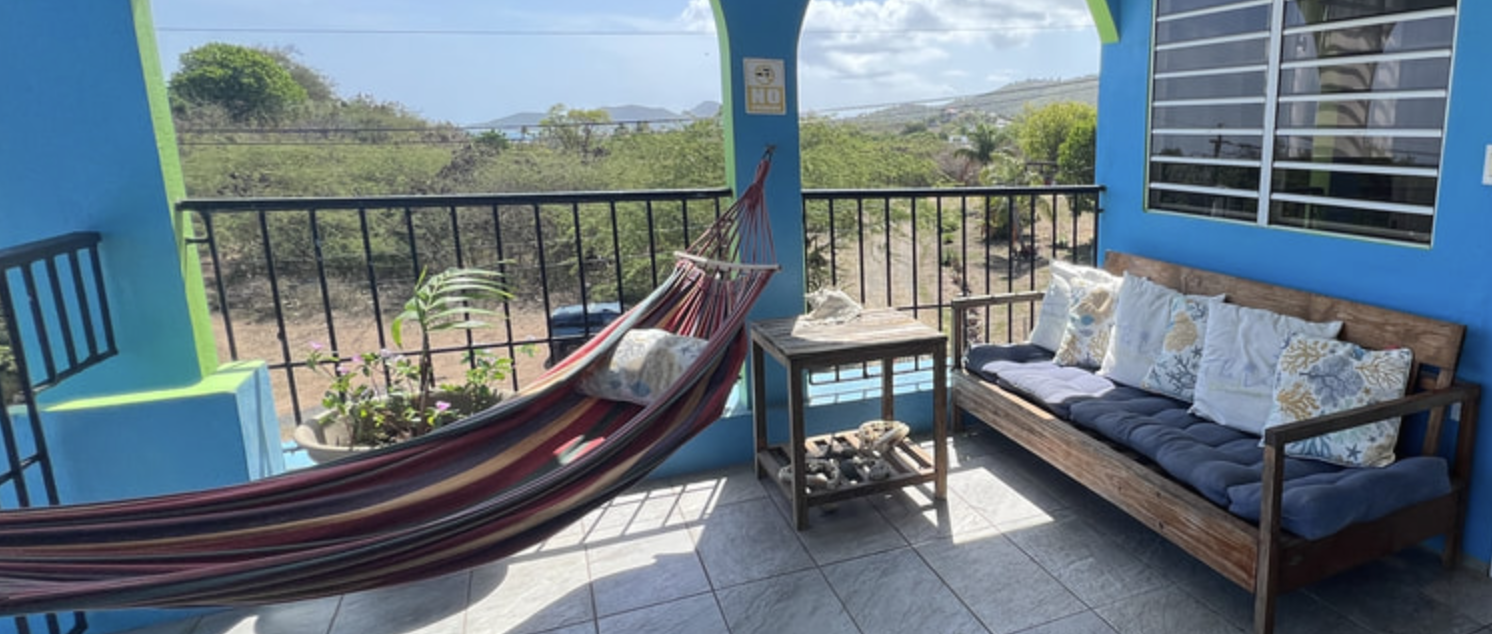 Balcony with a striped hammock, wooden bench with cushions, small table with decorative stones, potted plant, and view of trees and hills in the background.