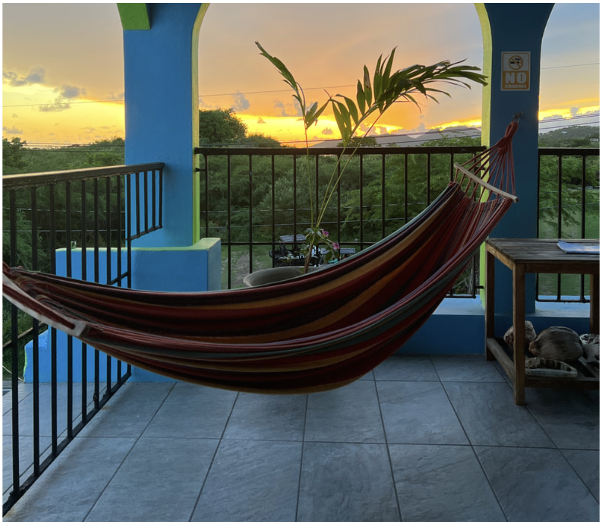 A colorful hammock on a balcony with a sunset view over green trees in the background. There is a small table with a plate and a plant in a pot, and a 'No Smoking' sign on the wall.