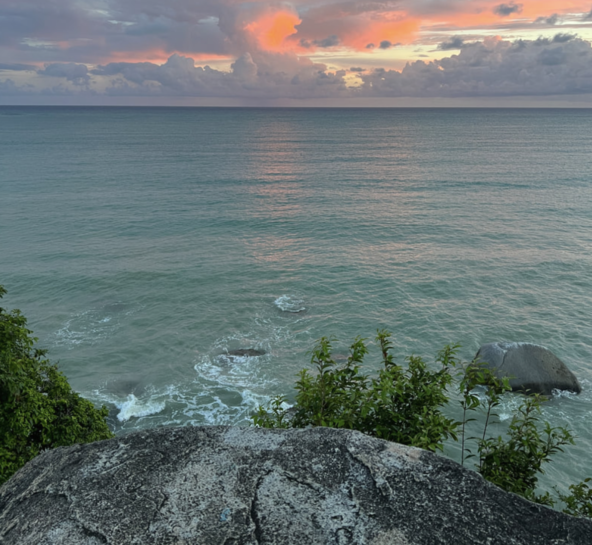 A seaside view at sunset showing calm ocean water, rocky shoreline with some greenery, and a colorful sky with pink and orange clouds.