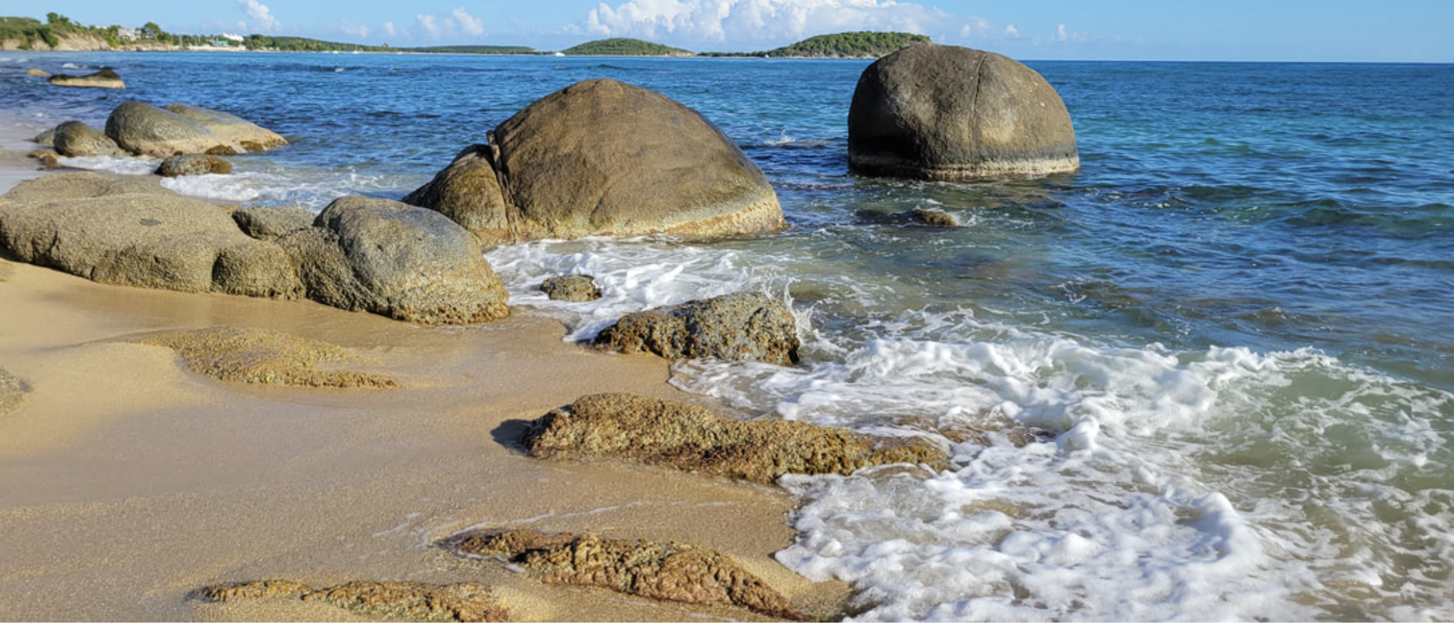 Sandy beach shoreline with large rocks, gentle waves, and distant green hills under a partly cloudy sky.