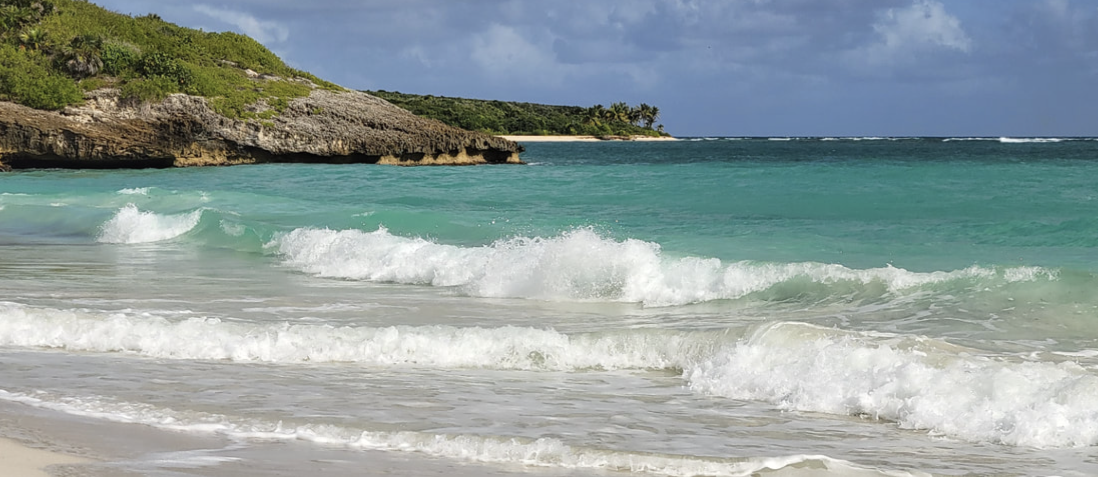 Turquoise ocean waves crashing on a sandy beach with a rocky cliff and palm trees in the background under a partly cloudy sky.