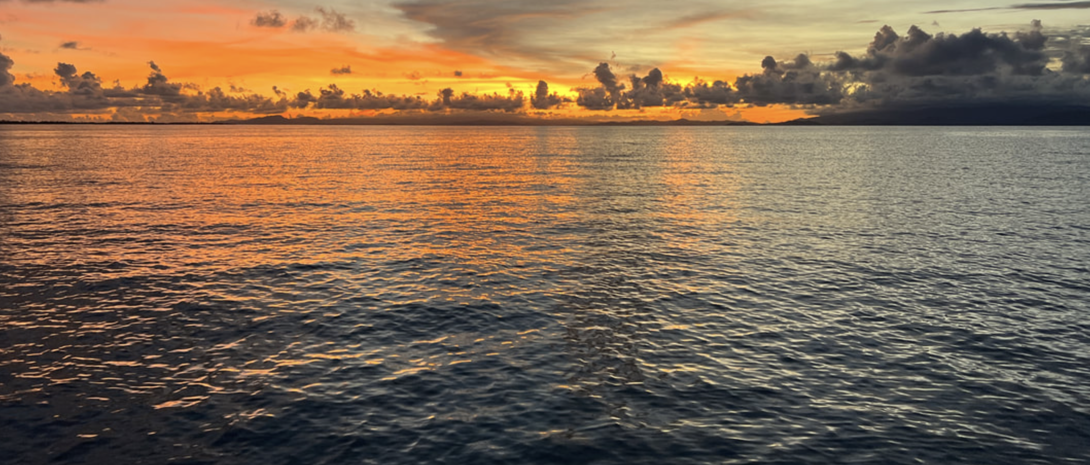 Sunset over the ocean with orange, yellow, and gray clouds in the sky and rippling water reflecting the colors.