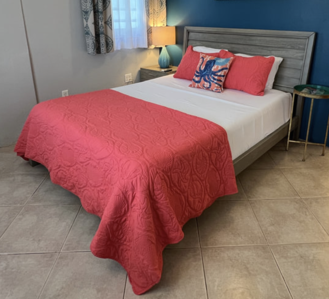 Hotel bedroom with a bed dressed in white sheets, a coral quilt, and three coral pillows. One decorative pillow features a blue octopus. There are curtains, a side table with a turquoise lamp, and a blue accent wall.