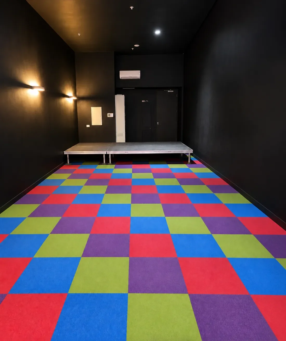 Workshop space at The Improv Pit in Collingwood, with a colourful checked carpet, black walls and a low stage at the far end.