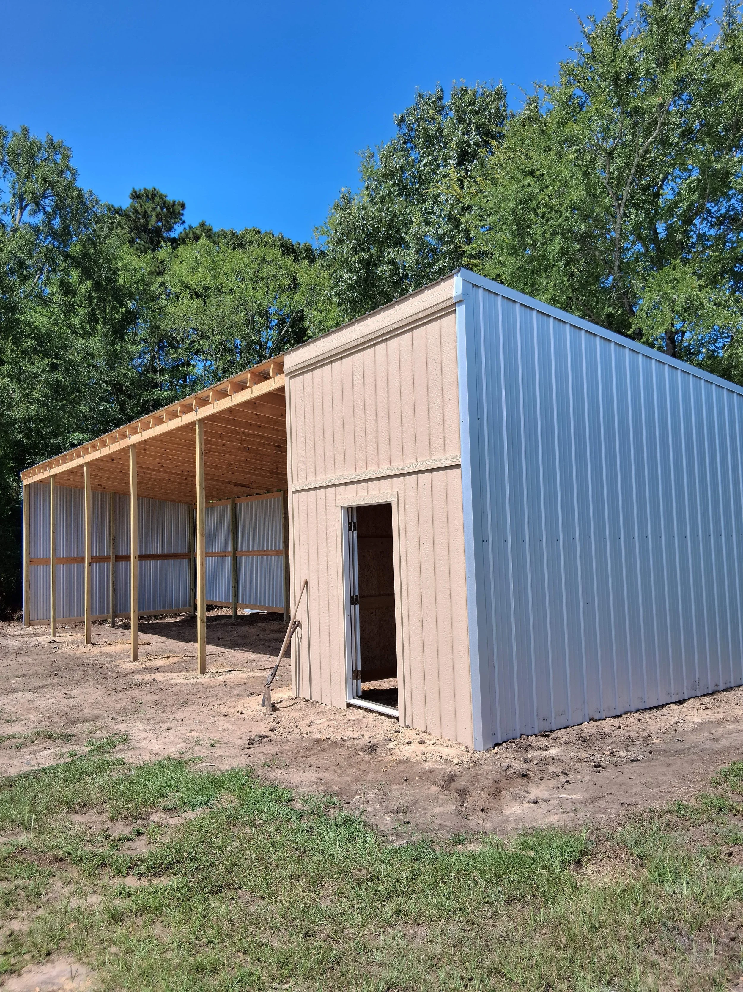 A building under construction with metal siding, wooden framing, and a partially built second level, surrounded by dirt and patches of grass, with green trees and a clear blue sky in the background.