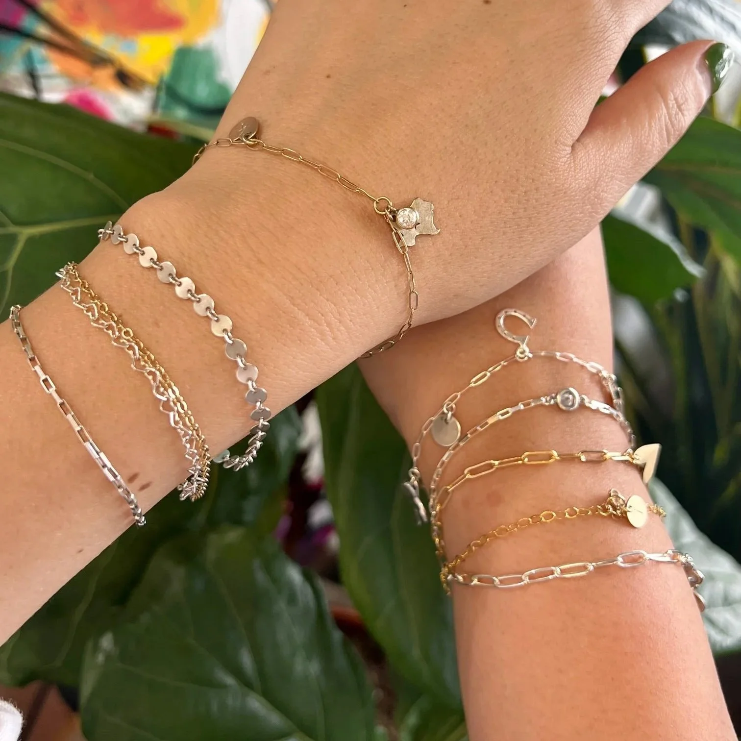 Close-up of a woman's arm and wrist with multiple gold and silver layered bracelets, against a backdrop of green leaves.