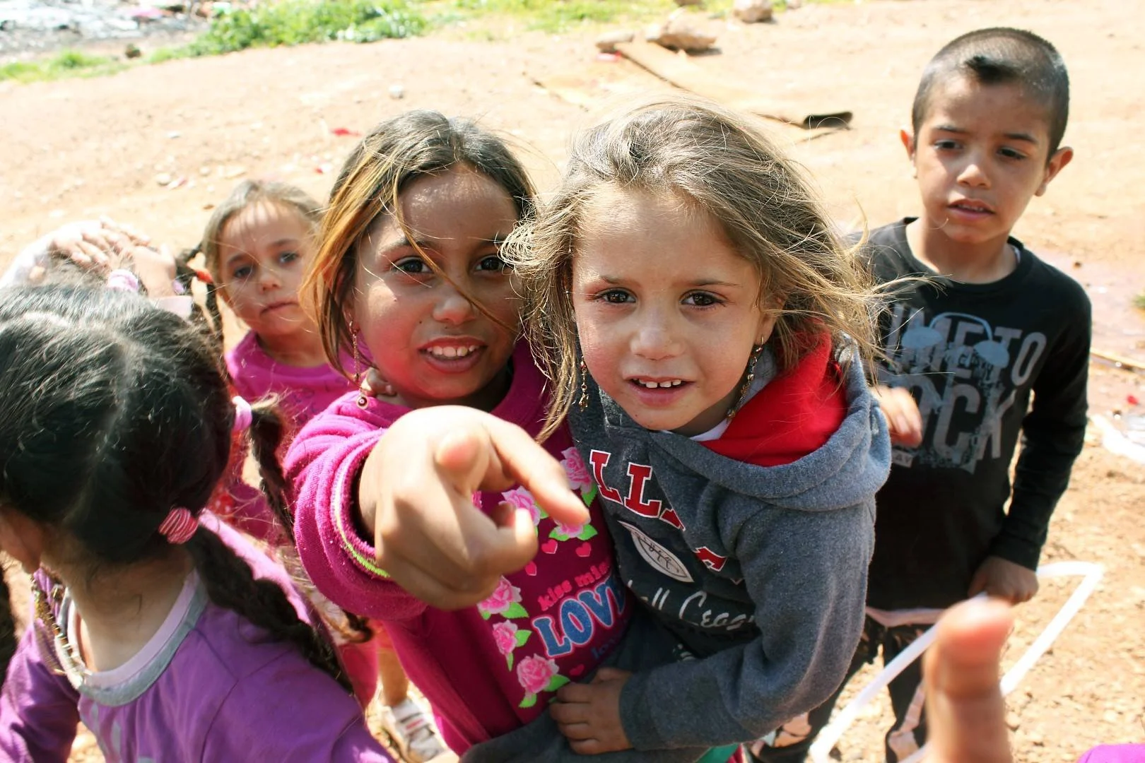 Group of young children outdoors, with one girl pointing at the camera, on a dirt ground with some plants in the background.