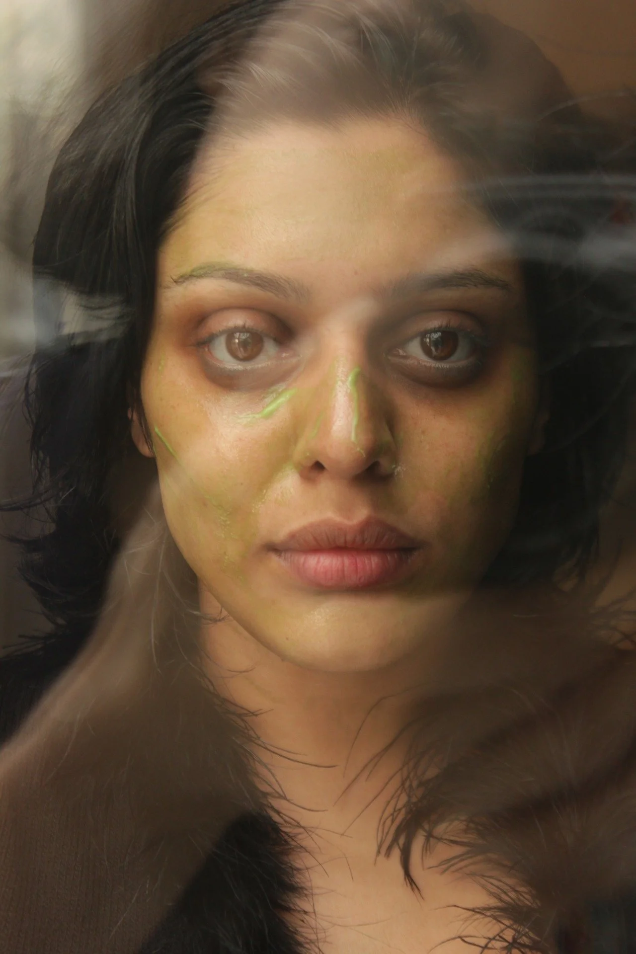Close-up of an artist with a green facial mask, dark hair, and brown eyes. There is a glass reflection creating a distorted effect.