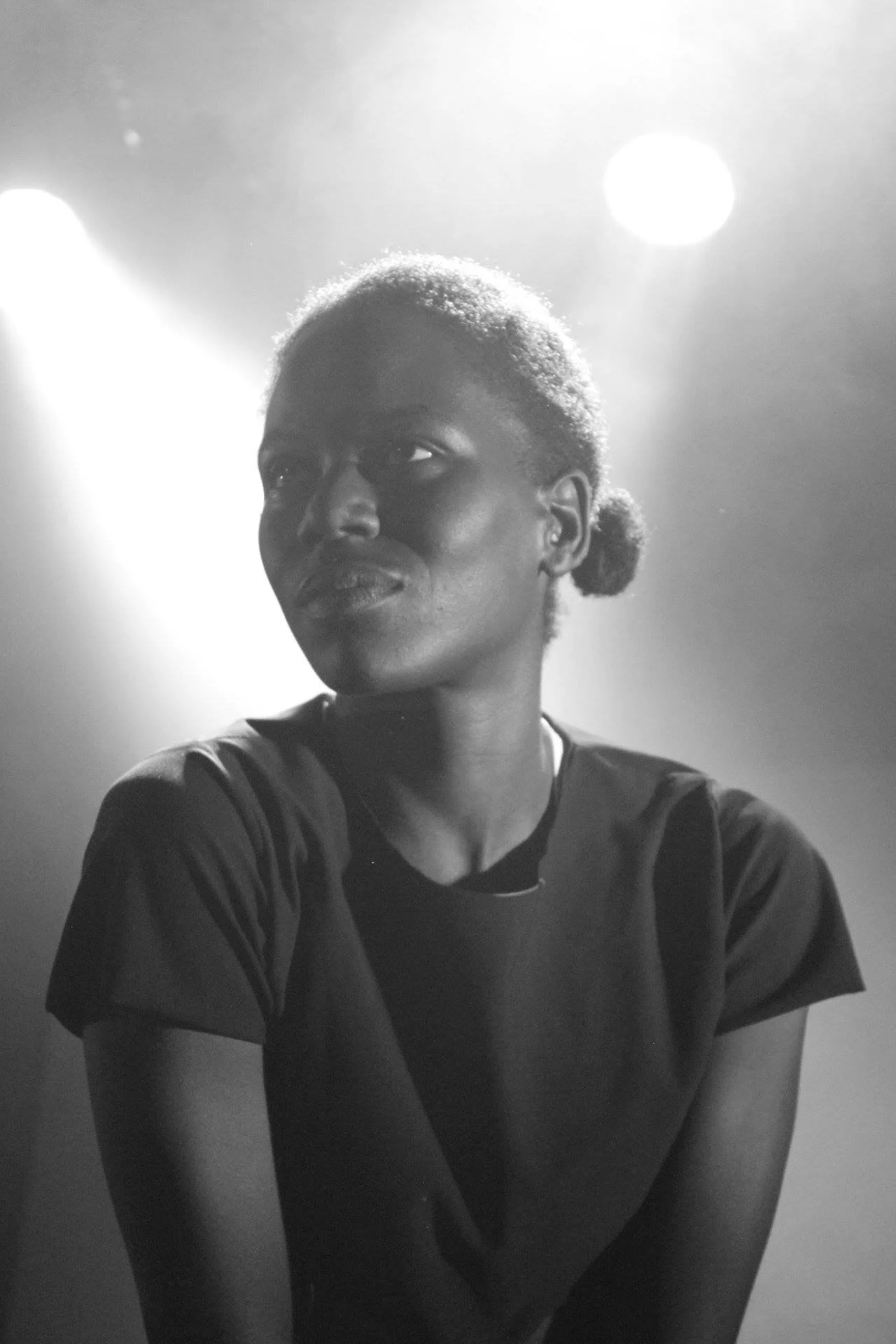 Black and white photo of a young black performer with short, curly hair, looking to the side with a slight smile, wearing a dark t-shirt, illuminated by bright light behind them.