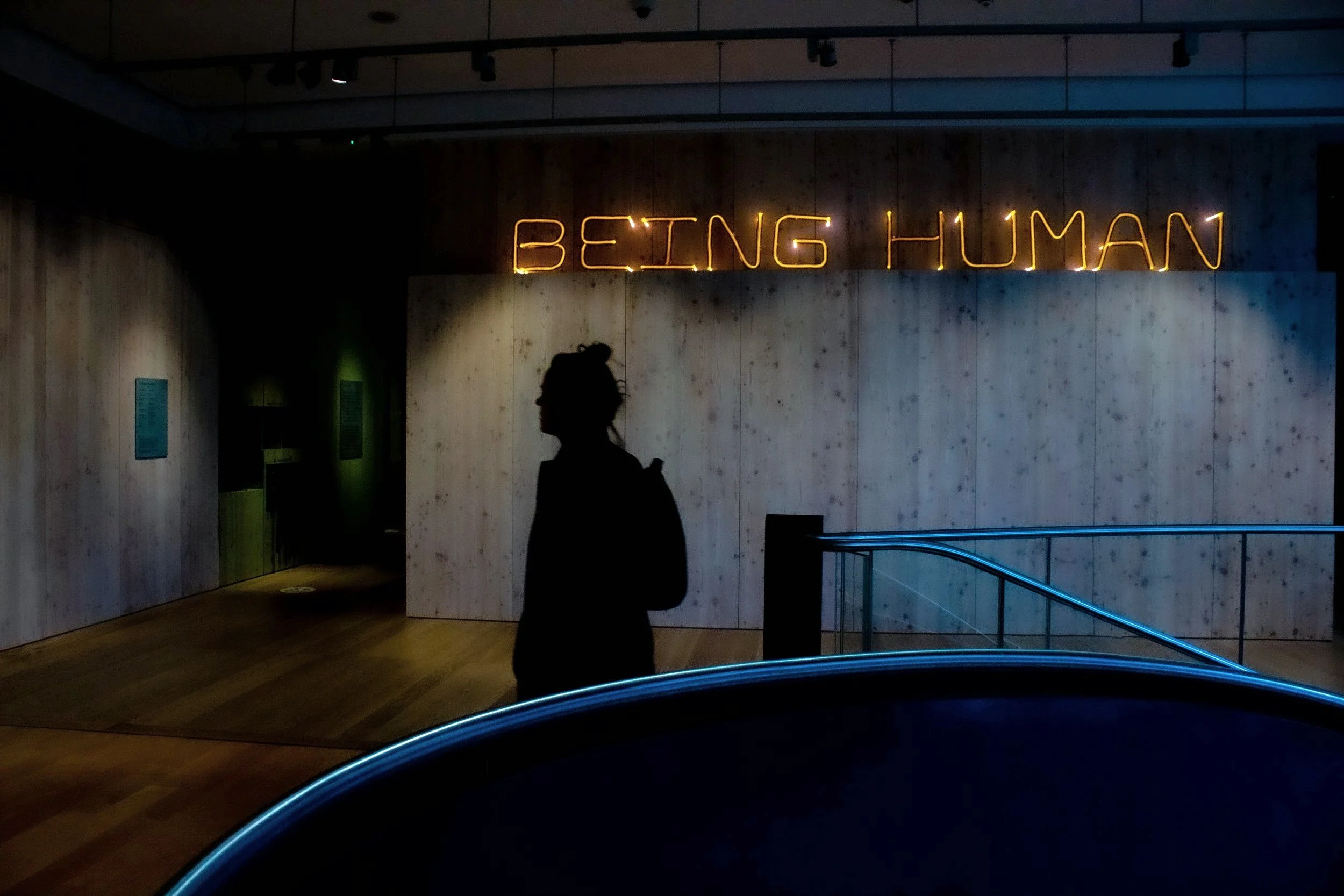 Silhouette of a person walking past a wall with neon sign that reads 'BEING HUMAN' inside a dimly lit space.