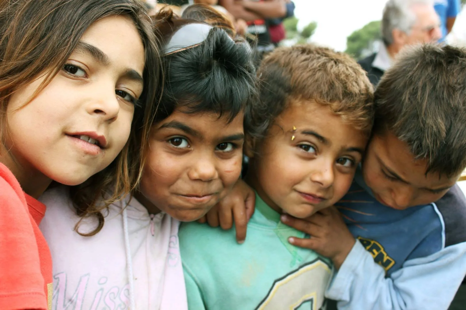 A group of four young children standing close together, looking at the camera, with a crowd blurred in the background.