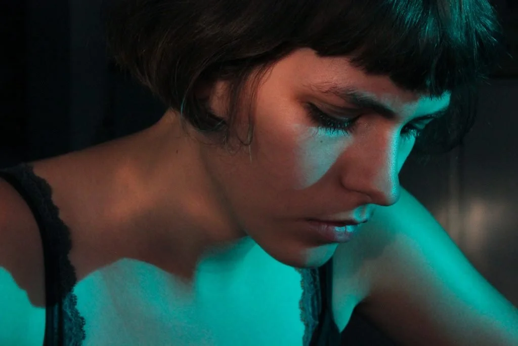 Close-up of a young woman with short dark hair, wearing a black lace top, looking down in a dimly lit environment with teal lighting.