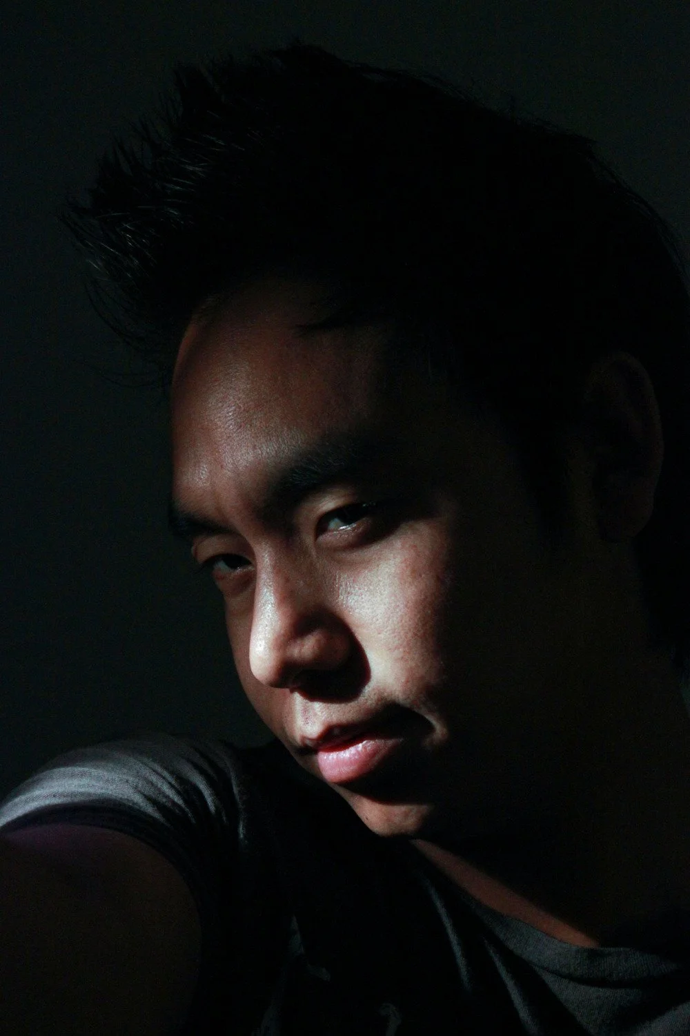 A close-up selfie of a young man with dark hair, illuminated by a soft light, against a dark background.