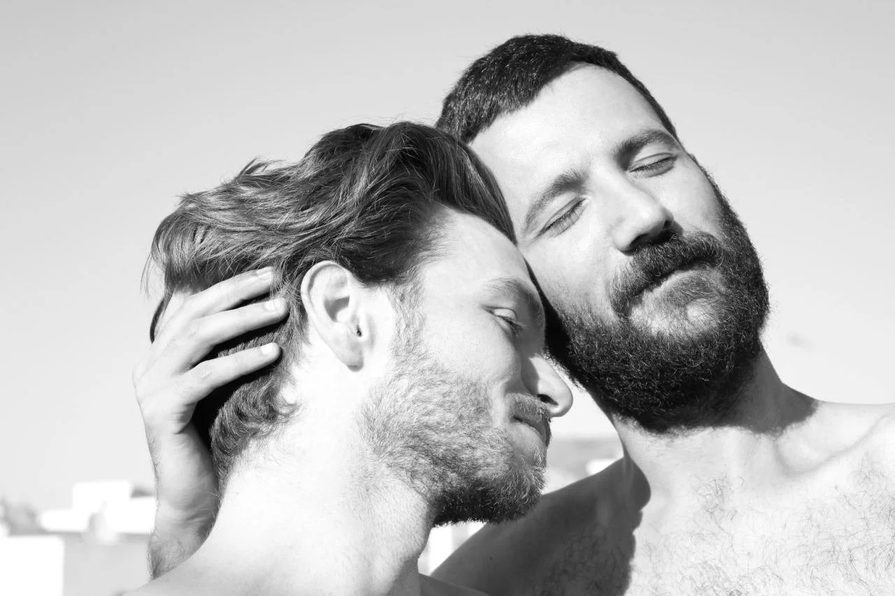 Two men with beards and closed eyes leaning their heads together, one resting his forehead on the other's head, in a close, intimate moment. Black and white photograph.