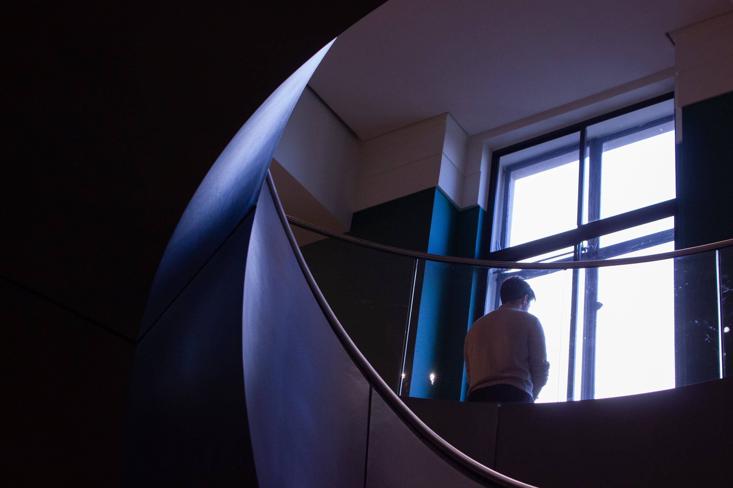 Interior view of a modern building staircase with a curved railing, large window letting in natural light, and a person standing near the window.