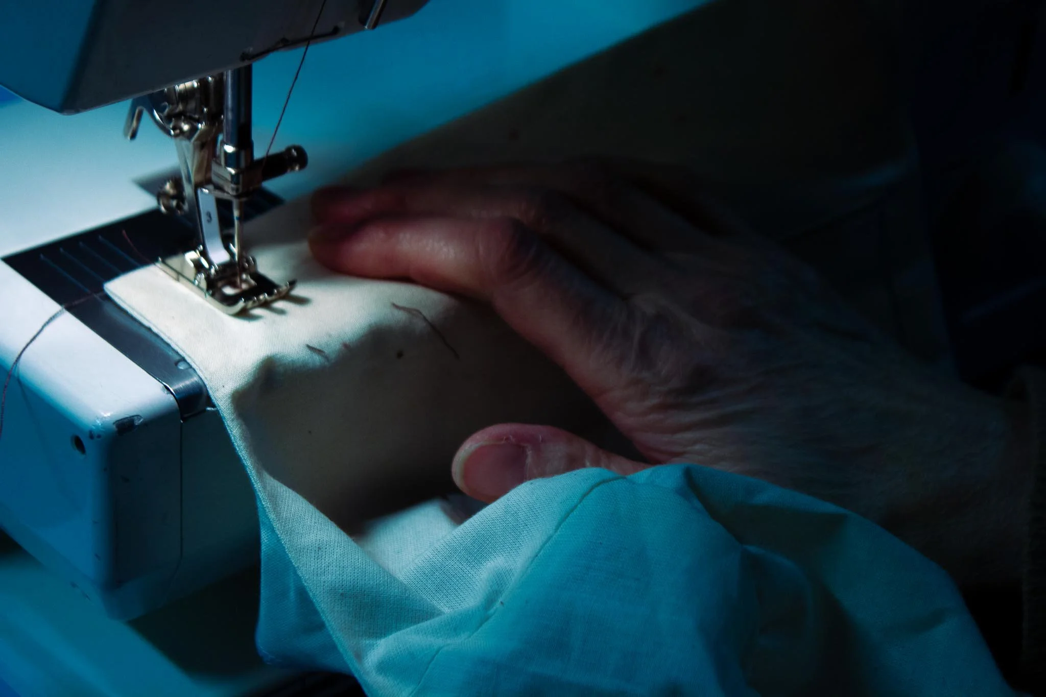 Close-up of a person's hand guiding fabric through a sewing machine in a dimly lit setting.