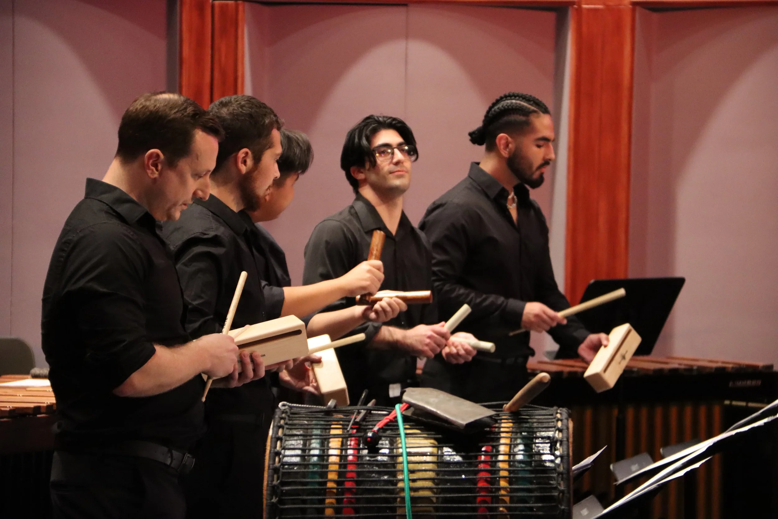 A group of five musicians dressed in black, holding percussion instruments, standing in music stance in a concert hall.