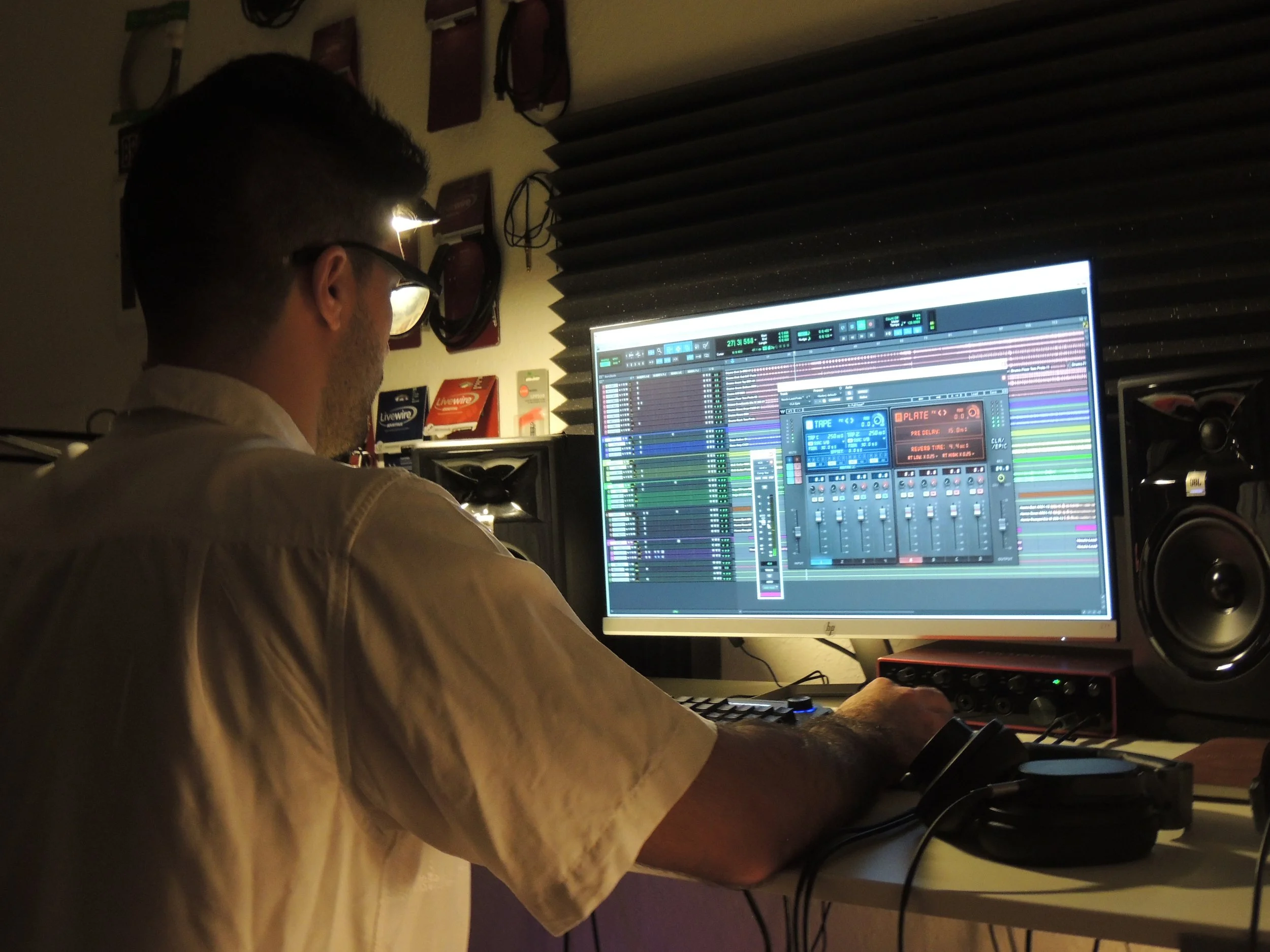A man working at a computer in a recording studio, with a music production software open on the screen, surrounded by audio equipment and soundproofing foam panels.