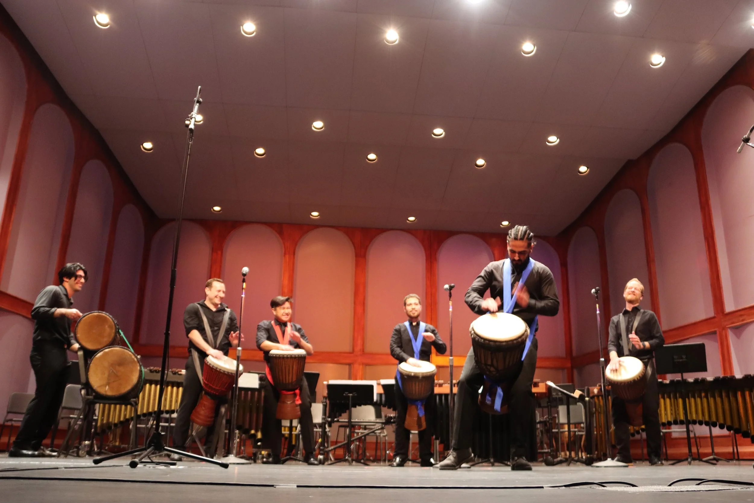 Group of seven musicians performing on stage with various percussion instruments, in a concert hall. They are dressed in black, and some are playing drums while others are clapping or using other percussion instruments.