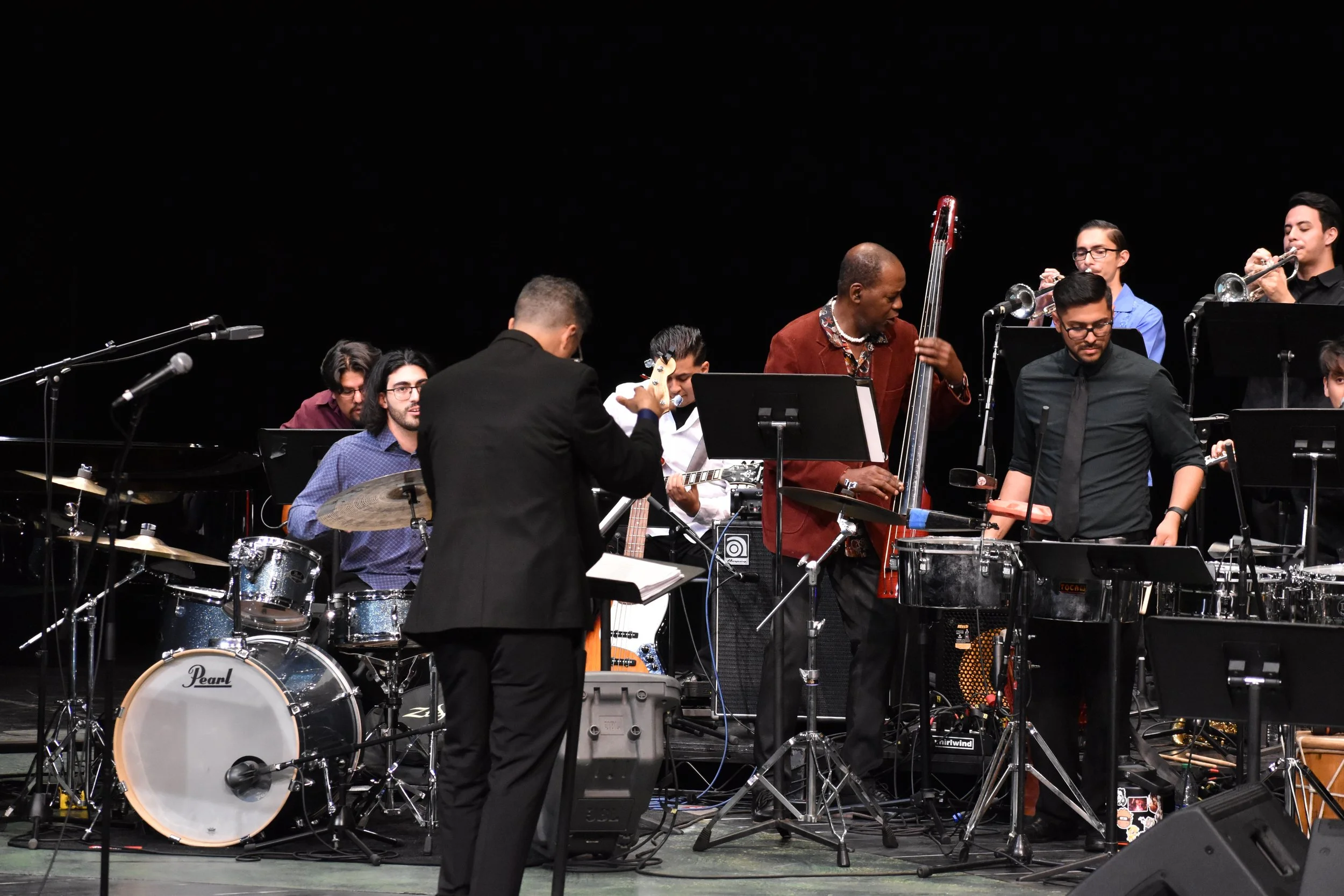 Jazz band performing on stage with musicians playing drums, trumpet, guitar, bass, and percussion.