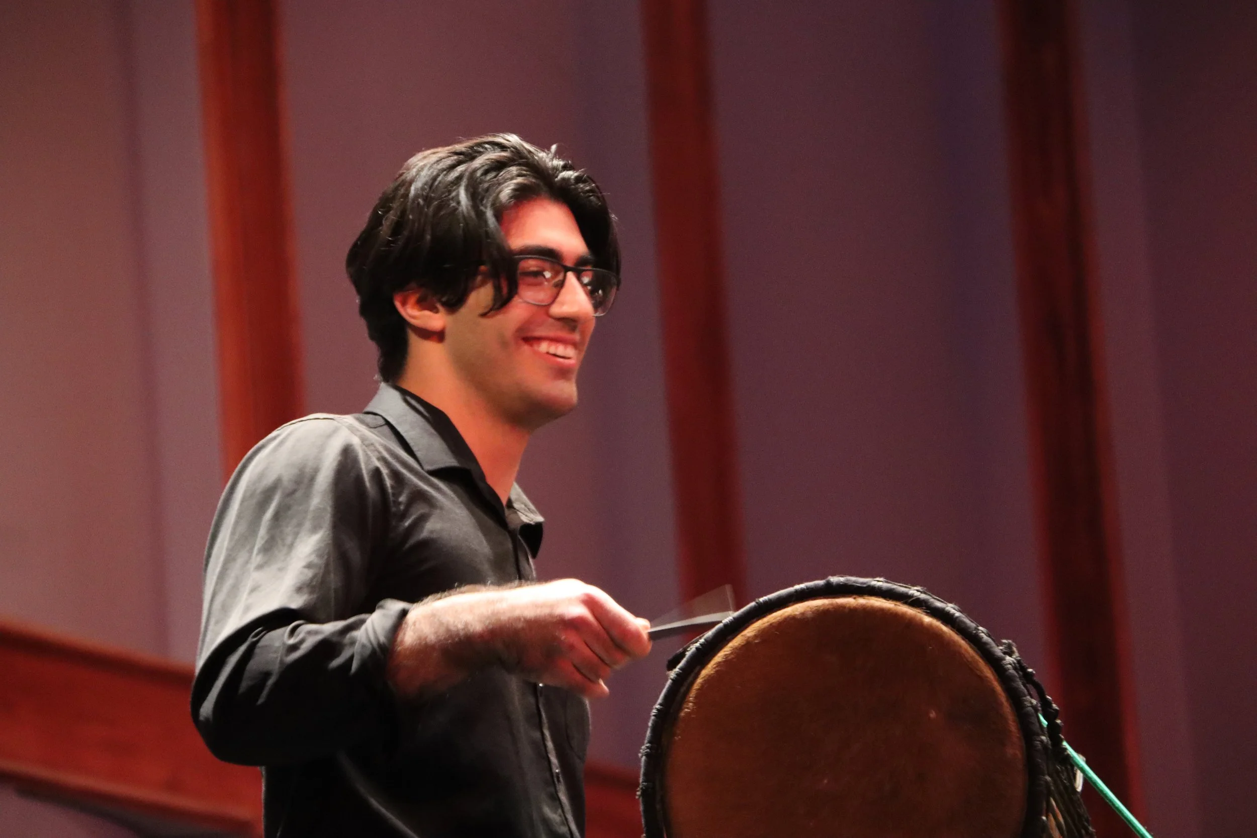 Young man with dark hair, glasses, and a black shirt smiling while playing a drum in a room with wooden paneling.