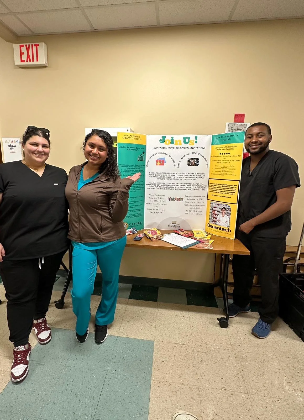 Three people in scrubs stand in front of a display board titled "Join Us!" with flyers and snacks on a table.