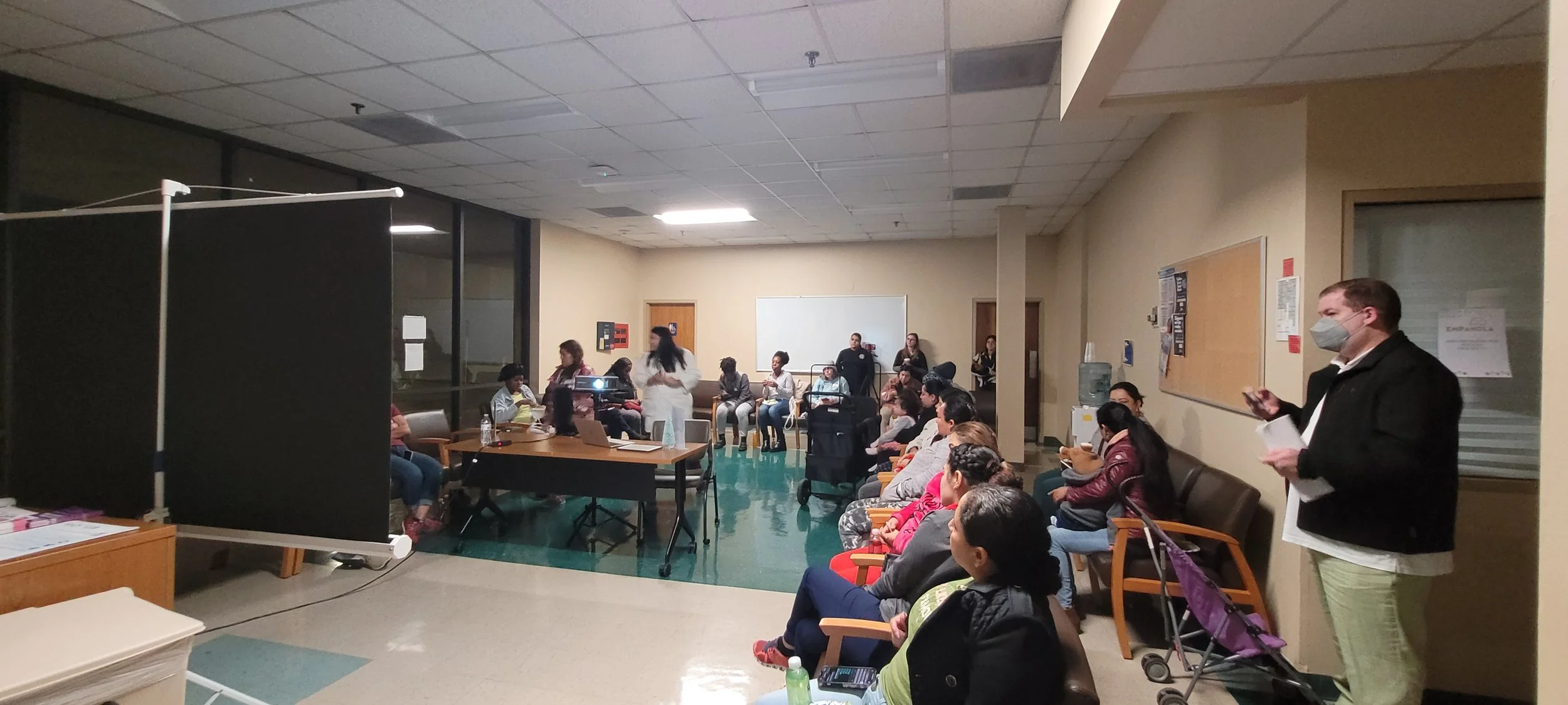 Group meeting in a room with people seated and standing, a table with a laptop, and a person speaking wearing a face mask.