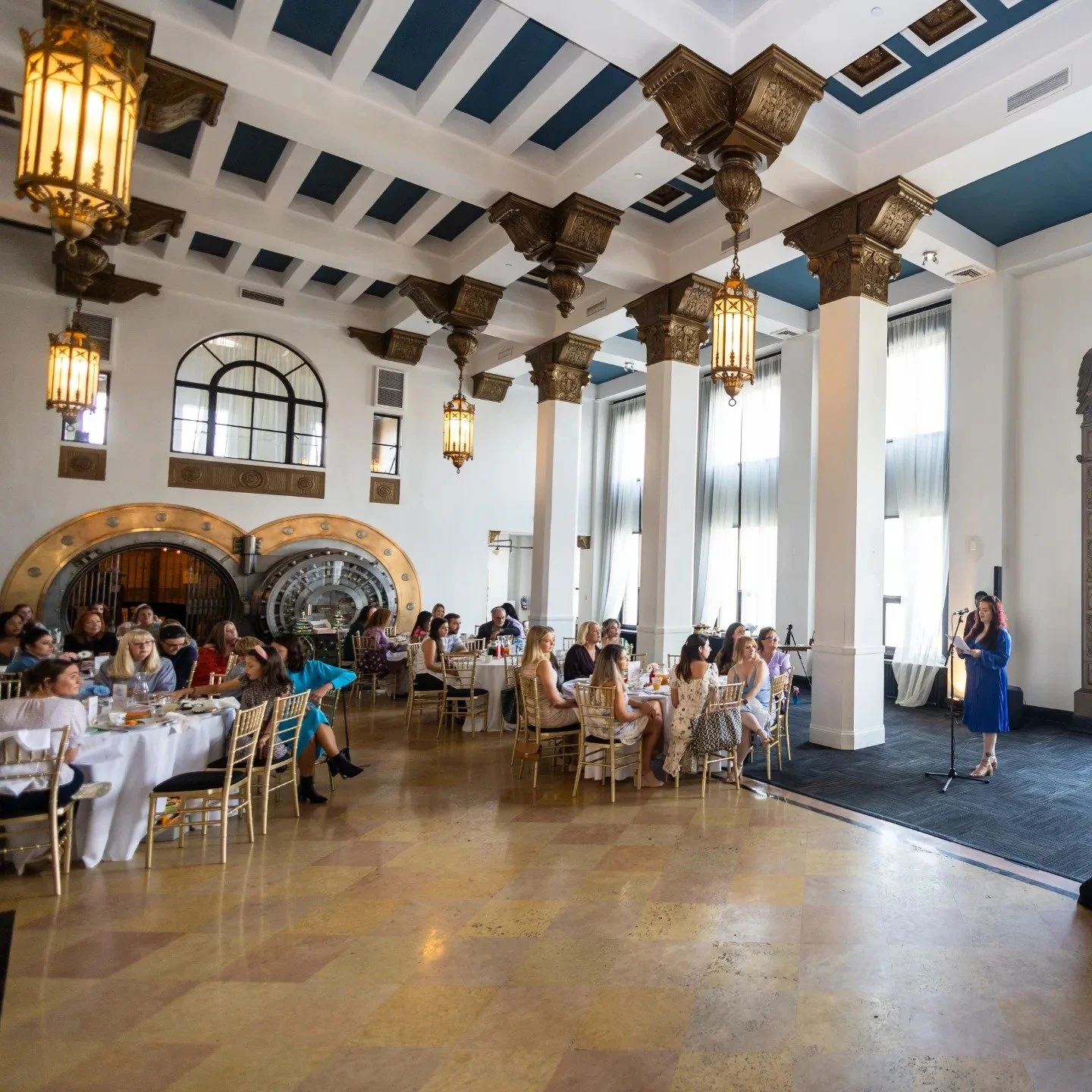 A large banquet hall with high ceilings, ornate columns, and tall windows with drapes. Several tables with white tablecloths are arranged, with guests seated. A woman in a blue dress is speaking at a microphone on a stage in front of the room.
