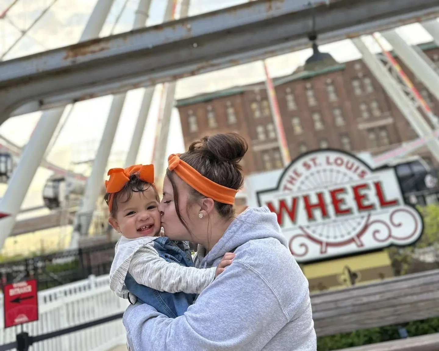 A woman holding a young girl at a fairground, with the 'The St. Louis Wheel' sign in the background.