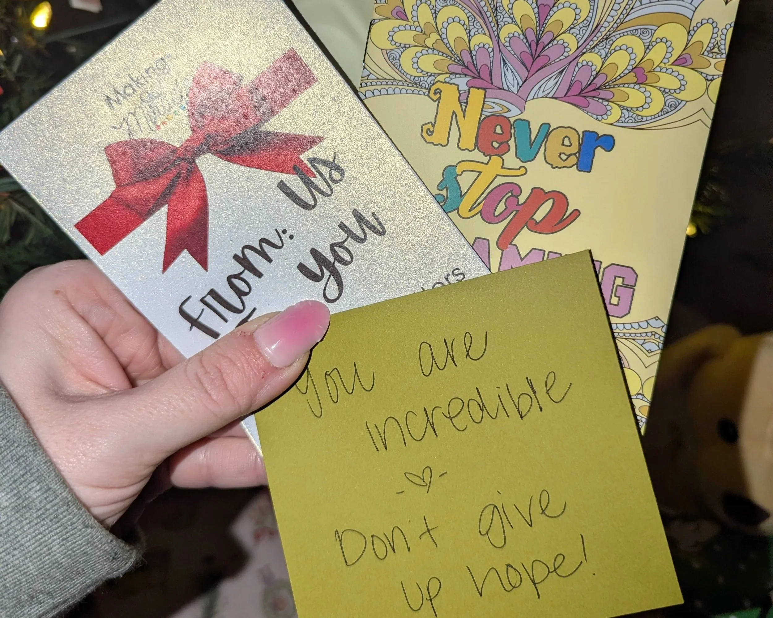 Close-up of a hand holding a shiny card with a pink and red bow illustration and text that reads 'A Mother is a Friend you can talk to about everything.' Behind the card, there is a colorful coloring page with the words 'Never Stop Being Amazing' and a decorative pattern on yellow paper.
