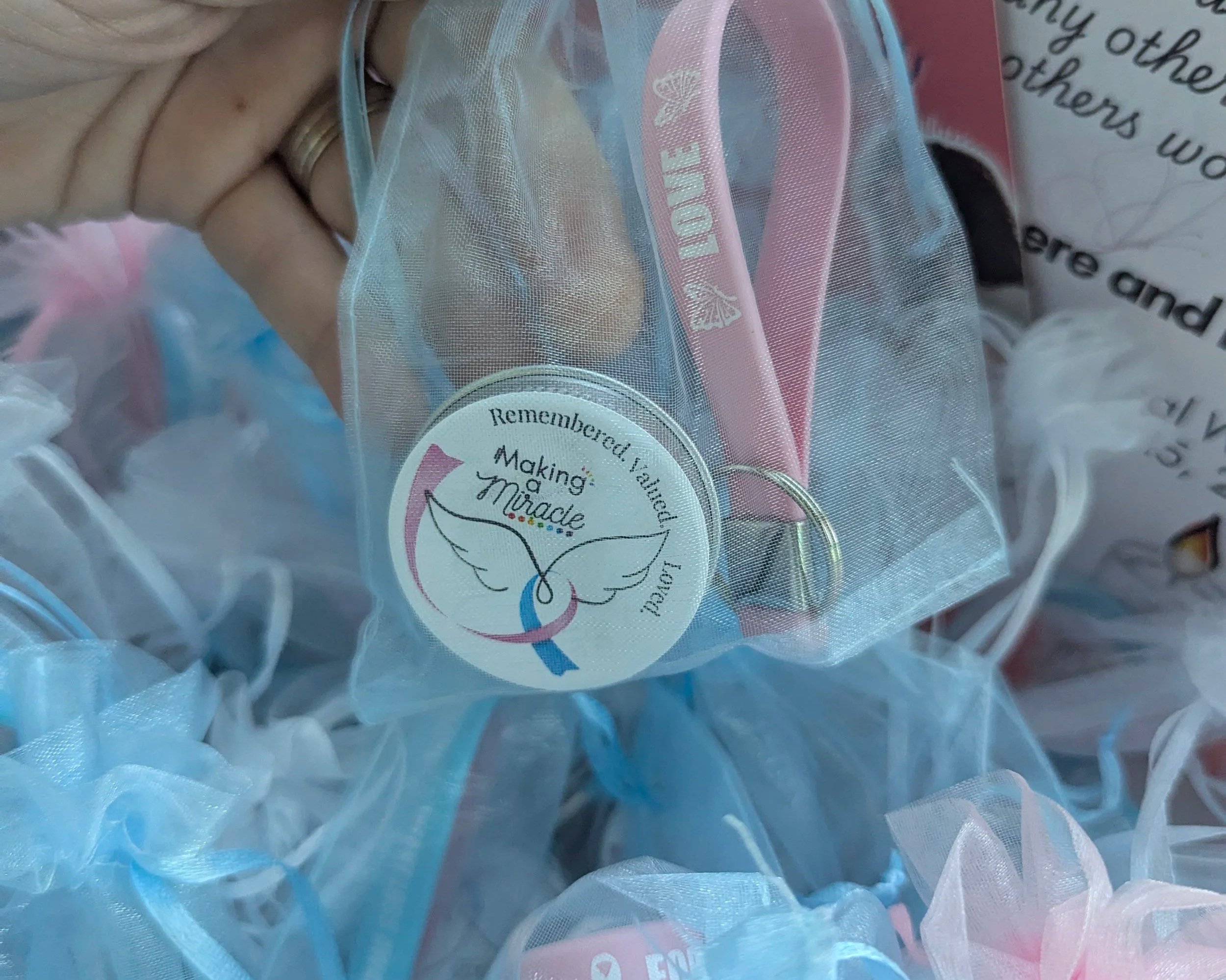 Close-up of a hand holding a mesh bag with buttons and a round tag that says 'Remembered... Making a Difference' with butterfly and floral illustrations, amid blue and white fabric.