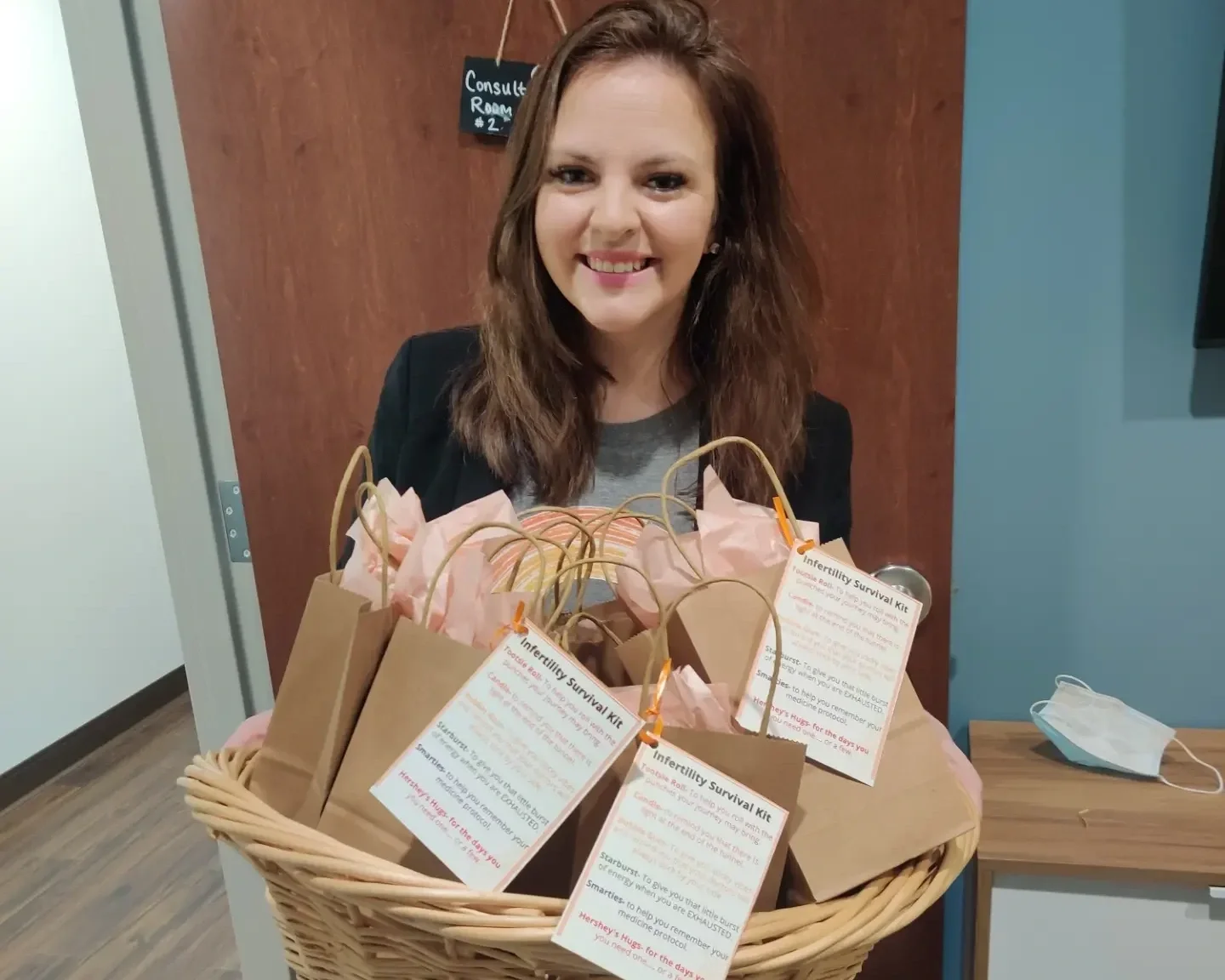 Smiling woman holding a basket with fertility survival kits and pink tissue paper, standing in a room with a wooden door and a blue wall.