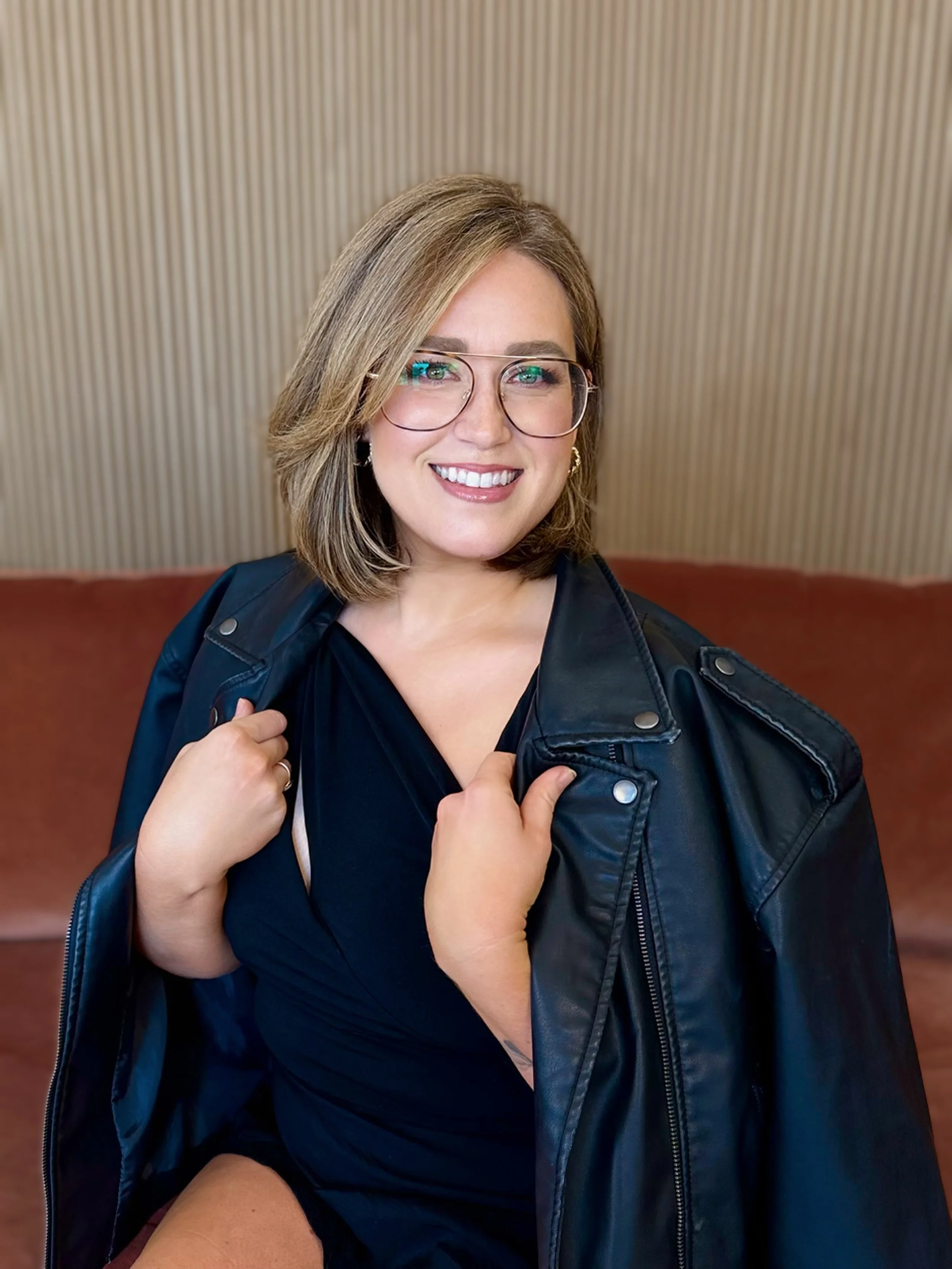 A woman smiling, wearing glasses, hoop earrings, a black lace top, and a dark jacket, sitting on a red couch with a wooden patterned wall in the background.
